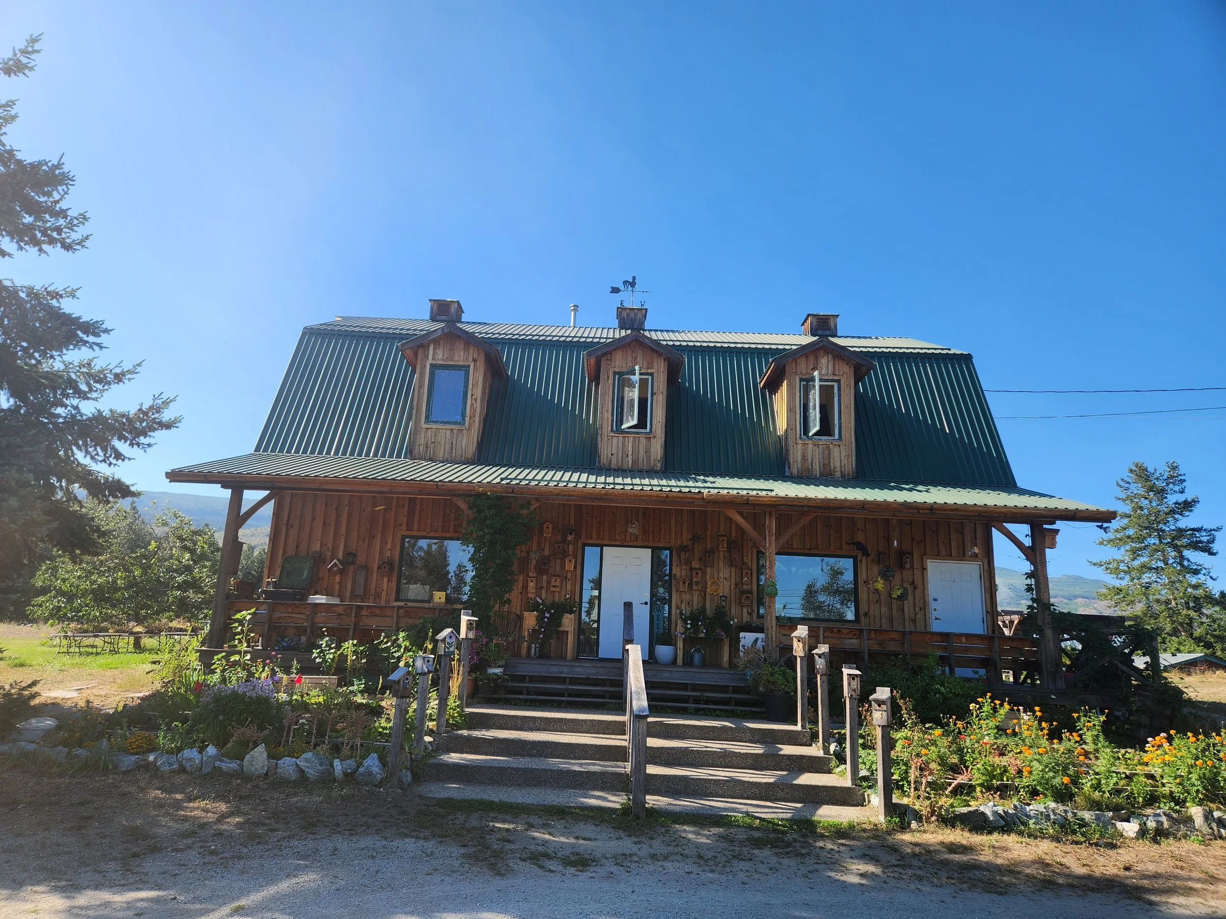 A two-story wooden house with a green metal roof and three dormer windows, surrounded by a garden with flowers and trees, under a clear blue sky.