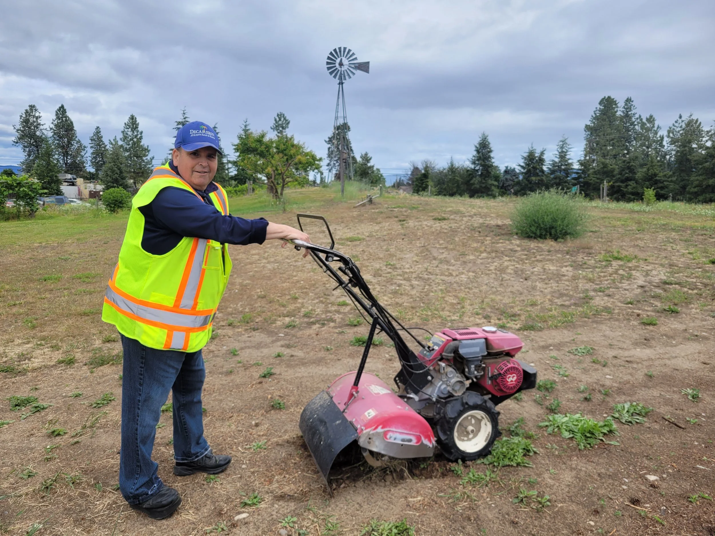 A person wearing a yellow safety vest and a blue cap is using a red and black dirt auger in a field with sparse green plants, trees, a windmill, and cloudy sky in the background.