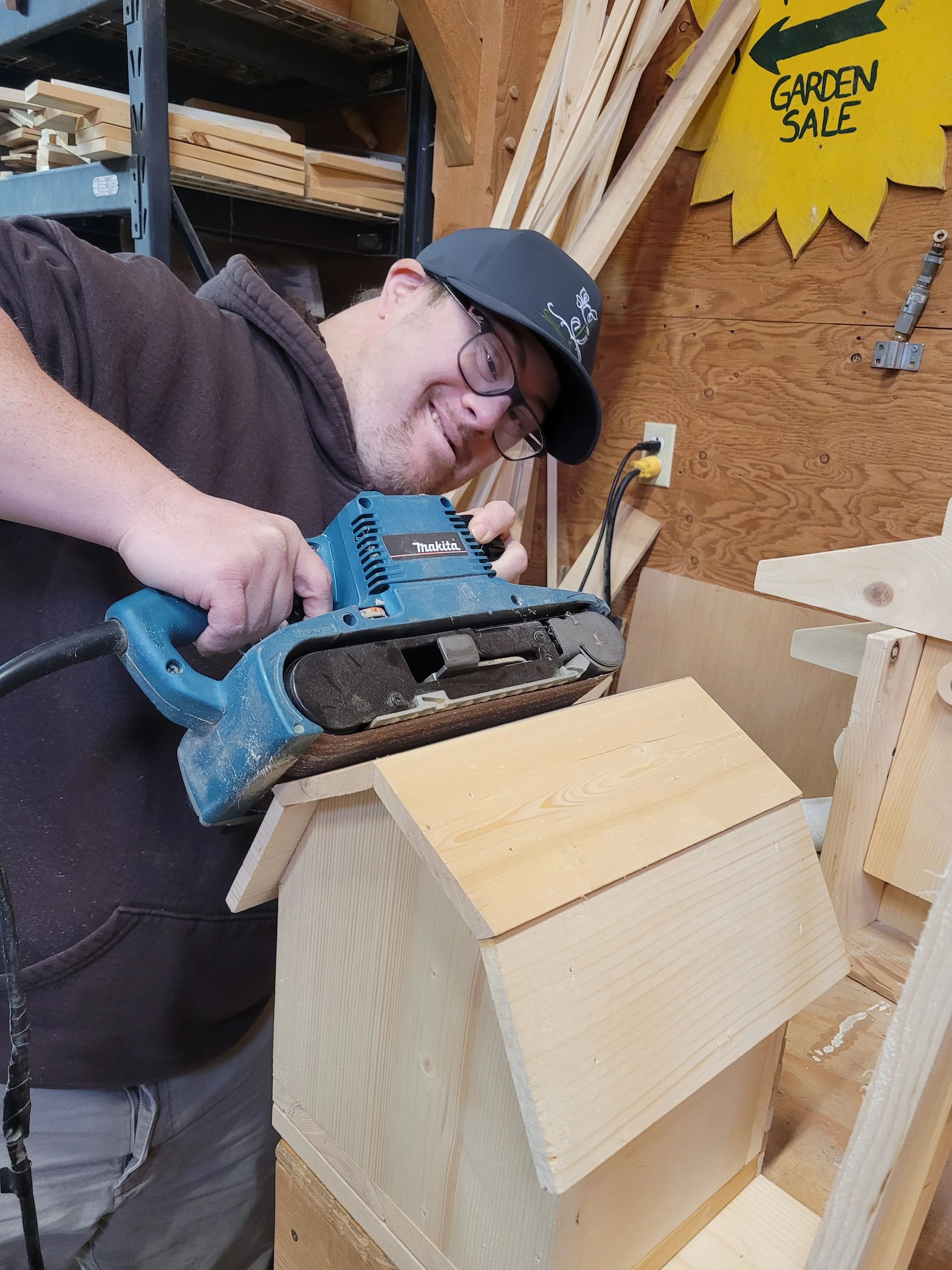 A man wearing glasses, a dark hoodie, and a baseball cap is using a handheld electric planer on a piece of wood in a woodworking workshop. The workshop background contains shelves with wood pieces, a yellow sign reading 'GARDEN SALE,' and wooden plan
