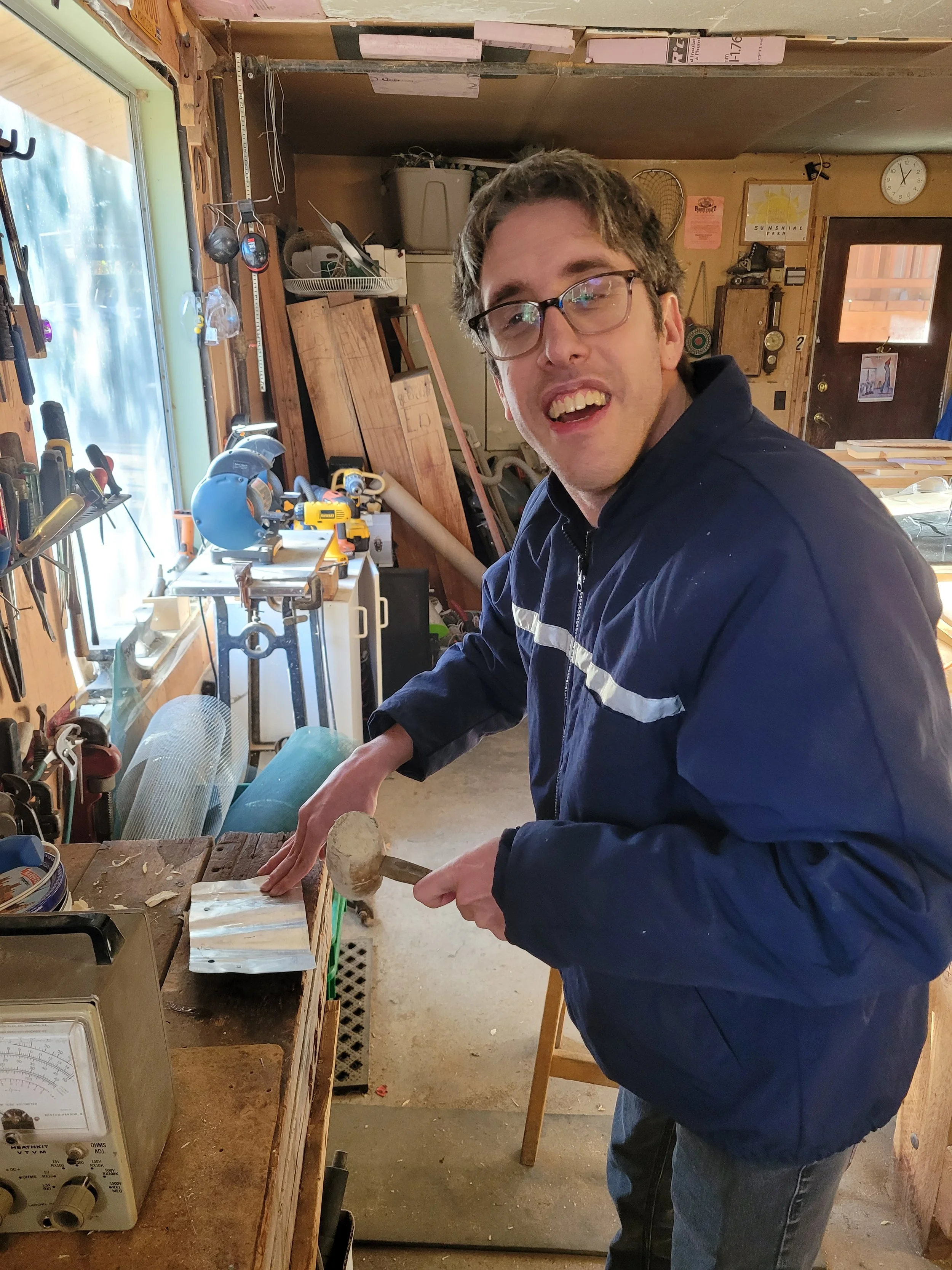 A young man in a workshop is holding a hammer and working on a piece of wood. The workshop has various tools on a wall, a scale on the table, and woodworking equipment in the background.
