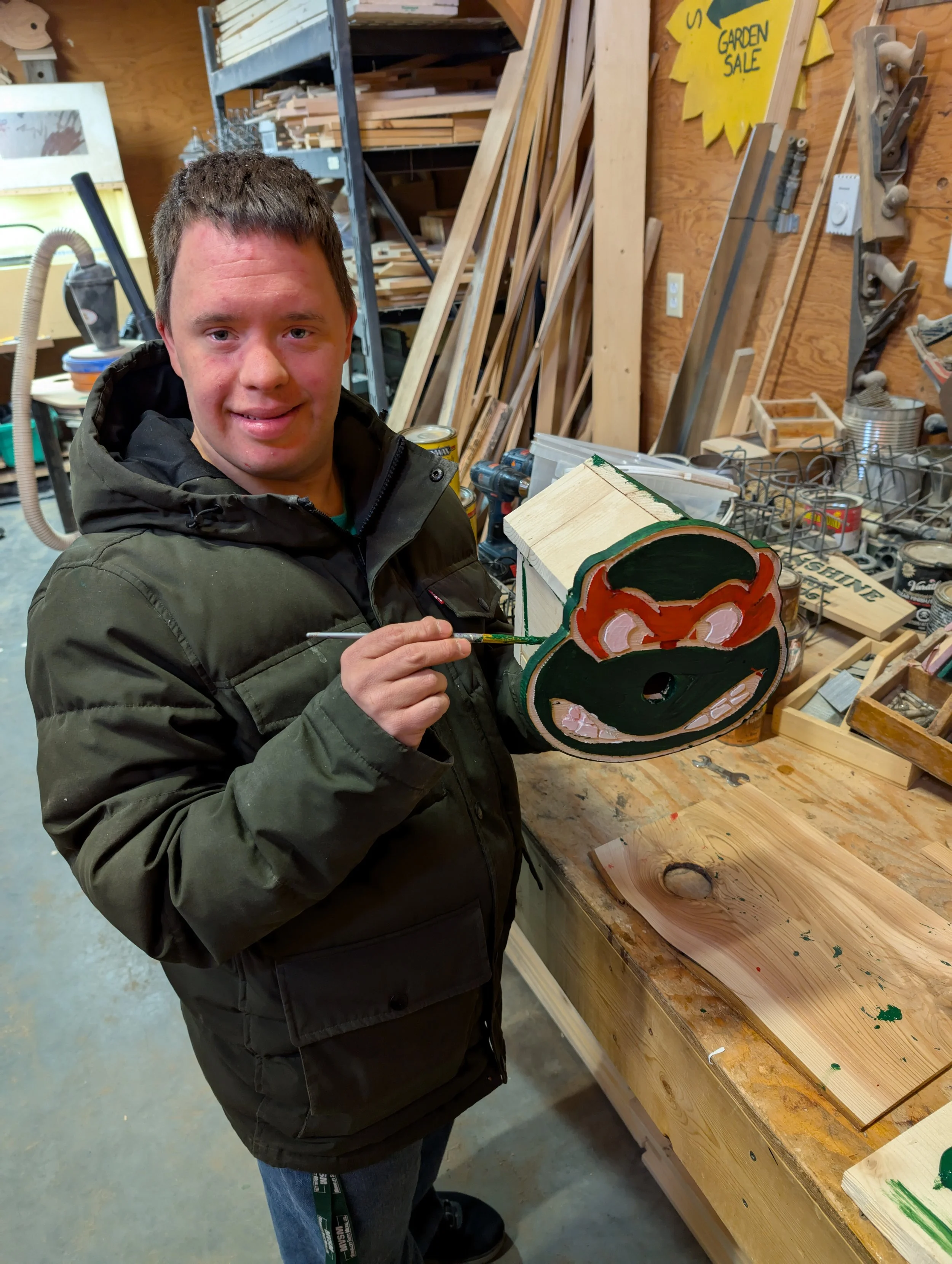 A man holding a painted wooden mask, standing in a woodworking workshop with tools, wood pieces, and a workbench around him.