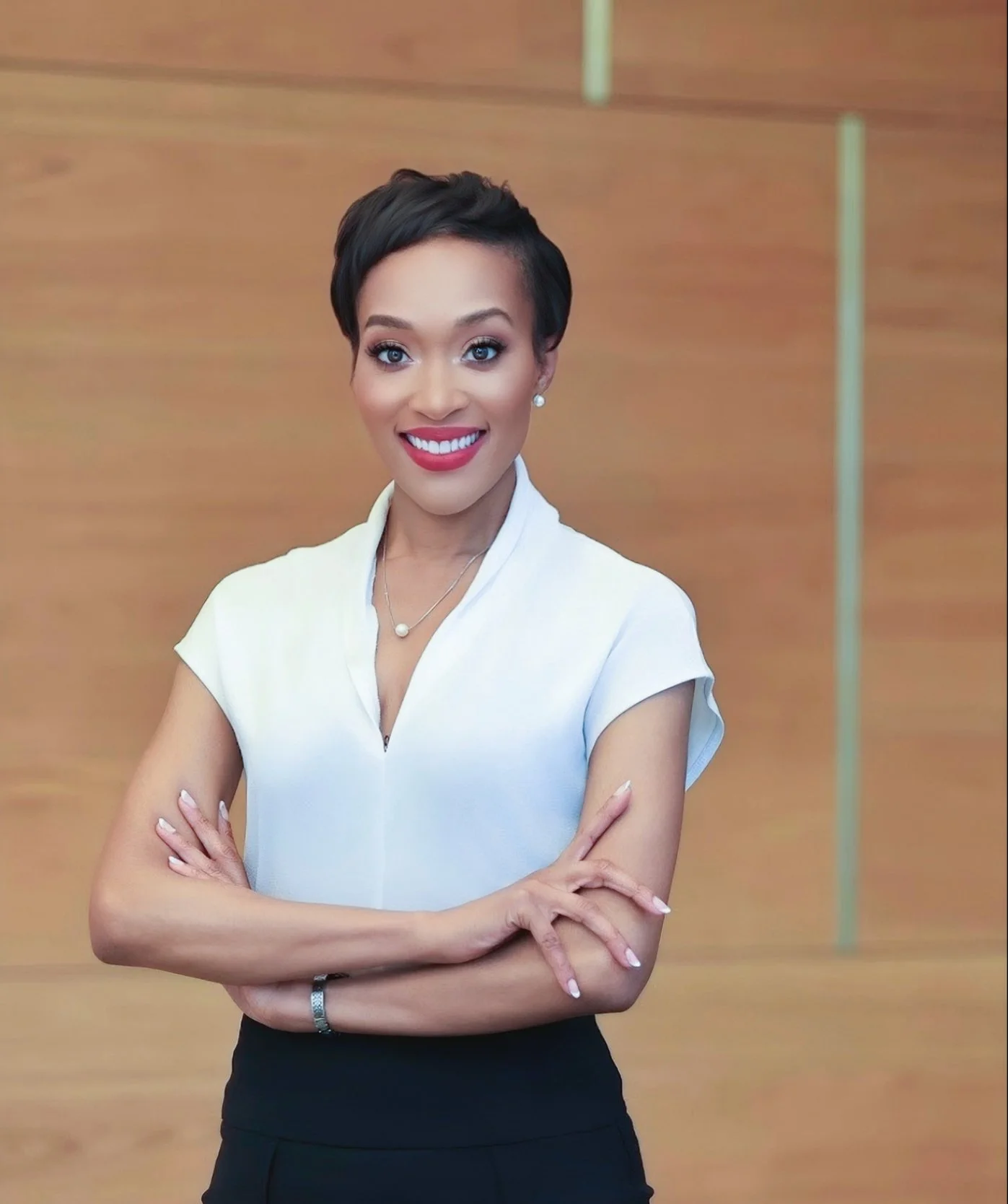 A smiling woman with short black hair, wearing a white shirt and black pants, standing with her arms crossed in front of a wooden background.