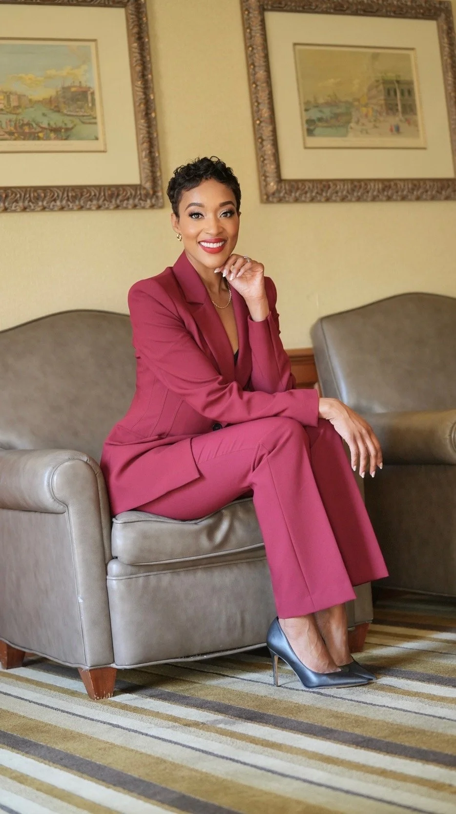 Professional woman with short black hair, wearing a black blazer and purple top, smiling with arms crossed in an office setting.