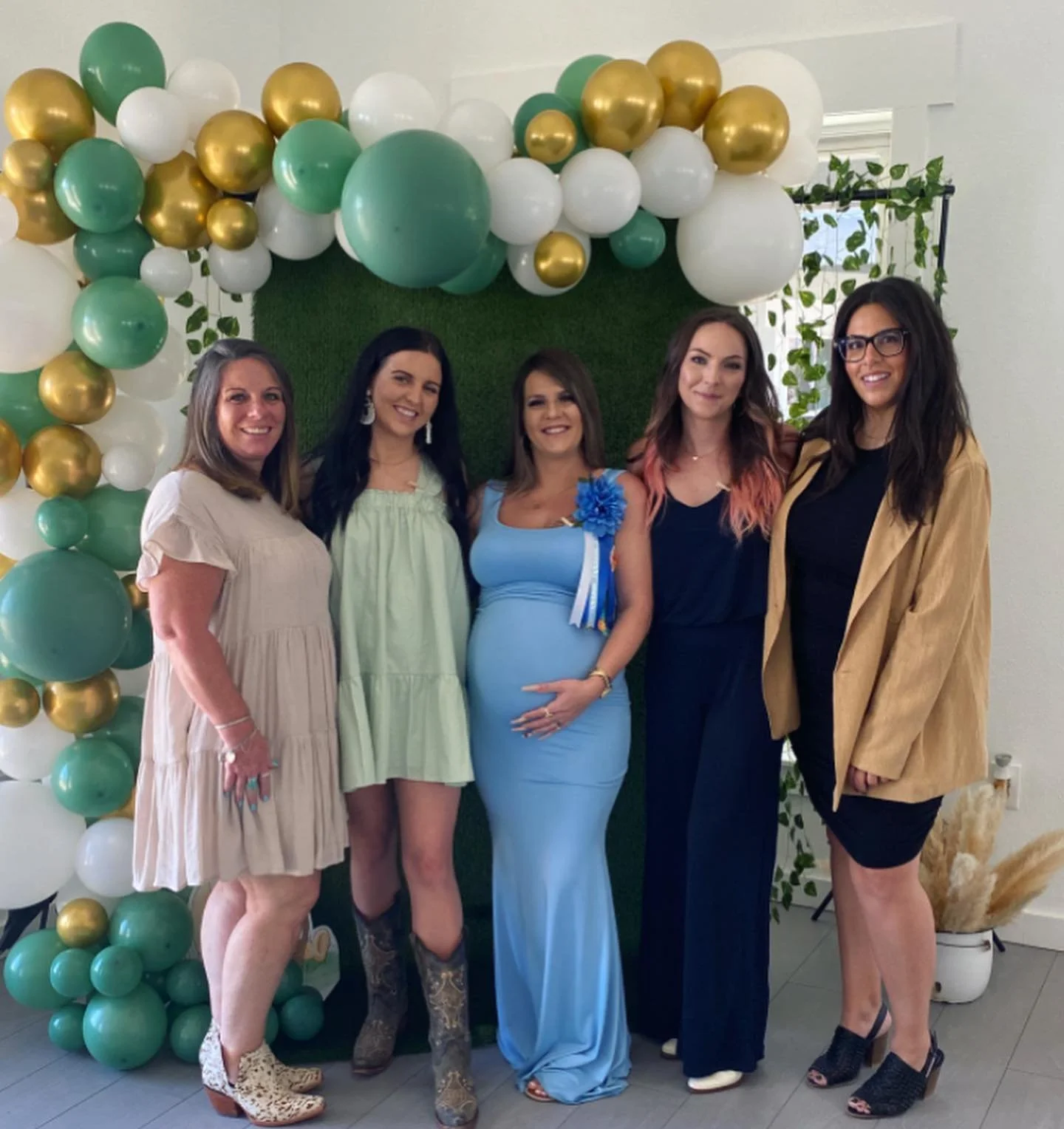 Five women standing in front of a colorful balloon arch and green backdrop, posing for a photo indoors.