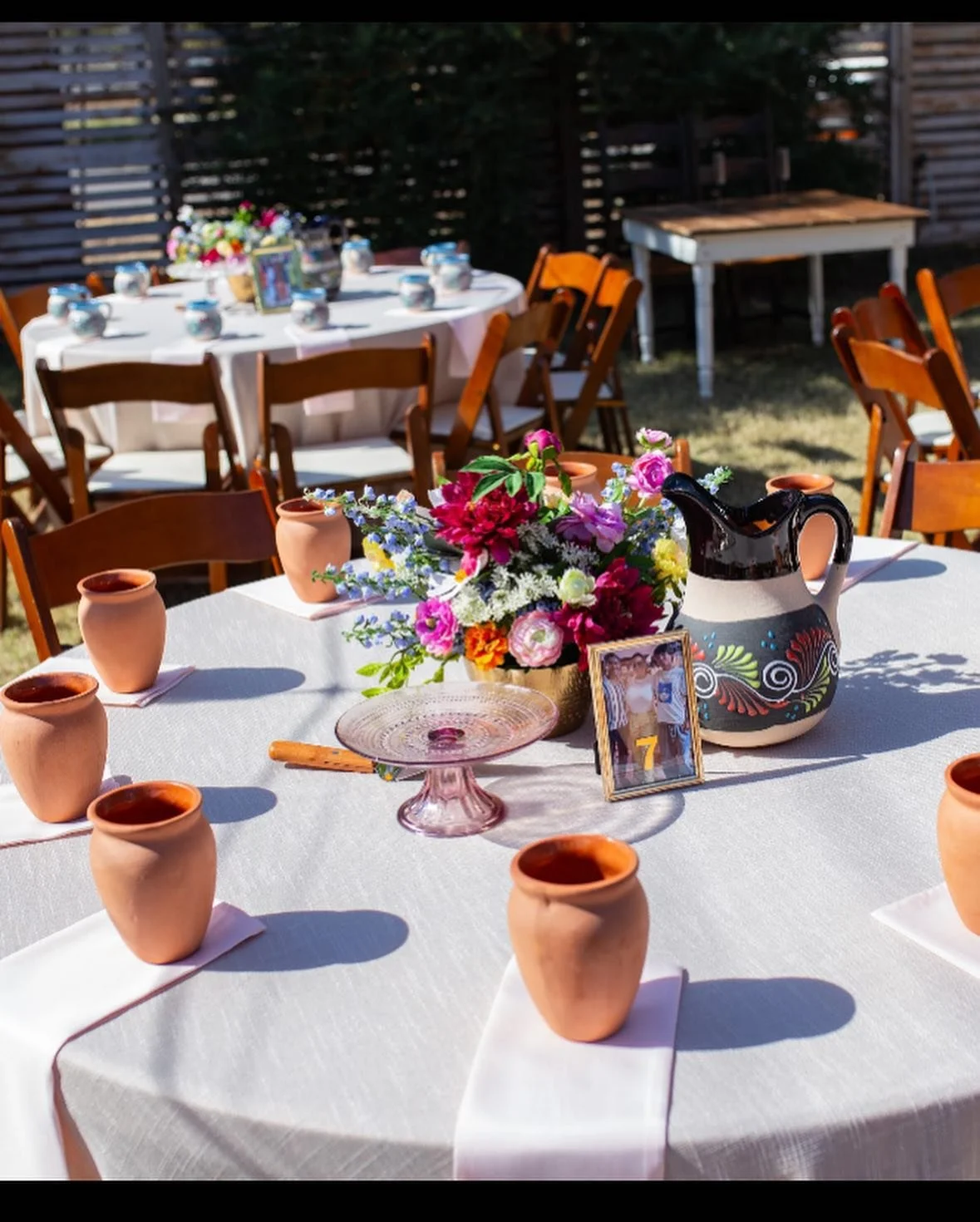A round table decorated for a celebration with a floral centerpiece, small terracotta jugs, a painted ceramic pitcher, a glass pink cake stand, and a photo frame. Other tables in the background have similar settings with chairs around them, set outdoors.