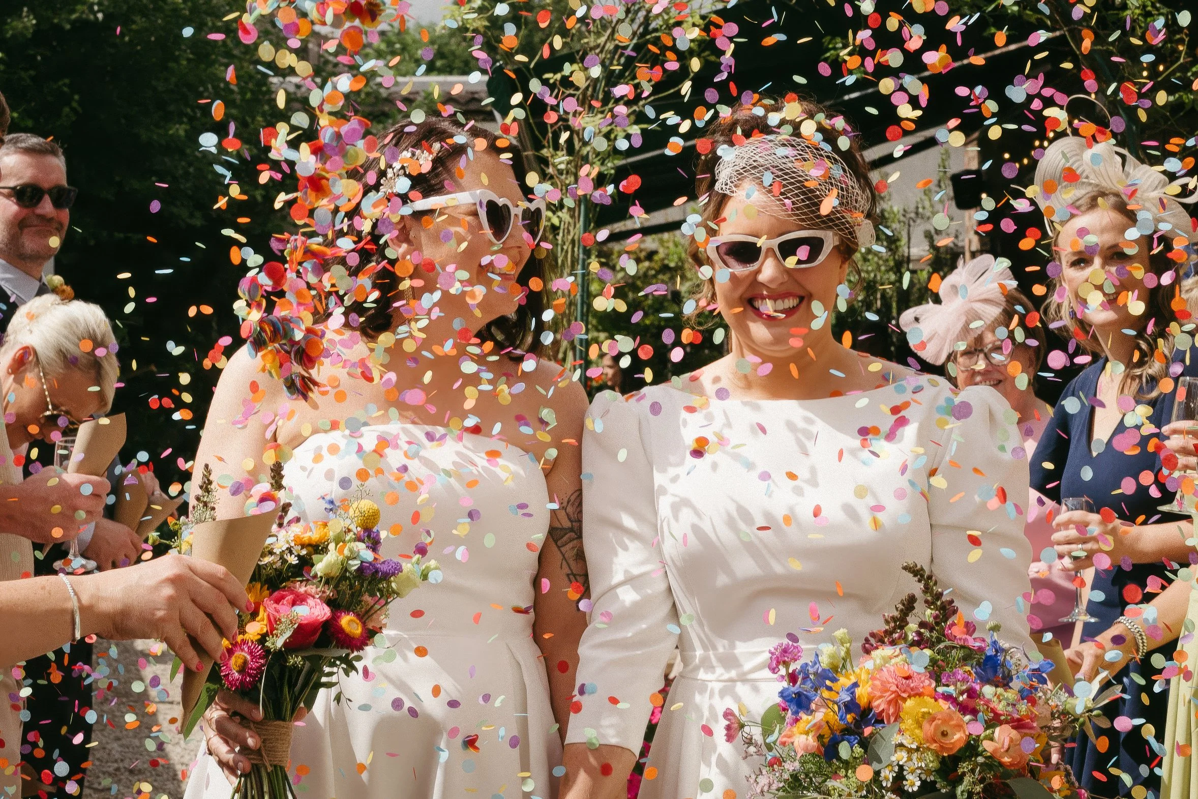 A group of women celebrating outdoors with confetti falling, dressed in white and colorful outfits, holding bouquets of flowers, smiling and wearing sunglasses. - captured by an Edinburgh wedding photographer