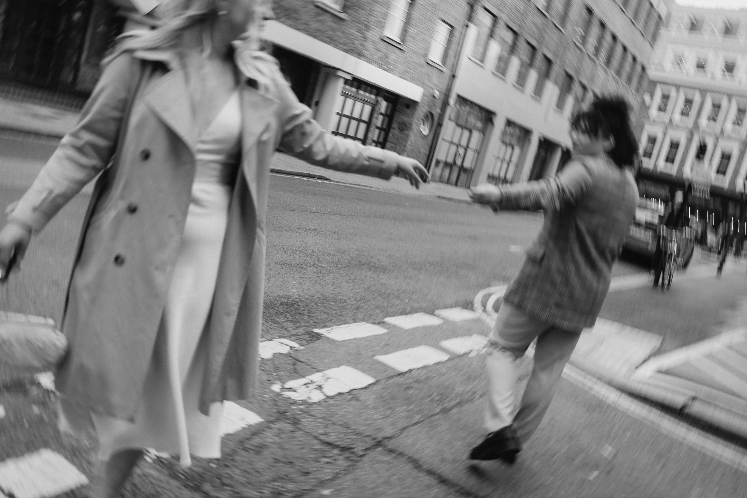 Two women on a city street, reaching hands toward each other as they walk past. One wears a trench coat, the other a checkered coat, both with long hair. The surroundings include buildings, parked cars, and a pedestrian.