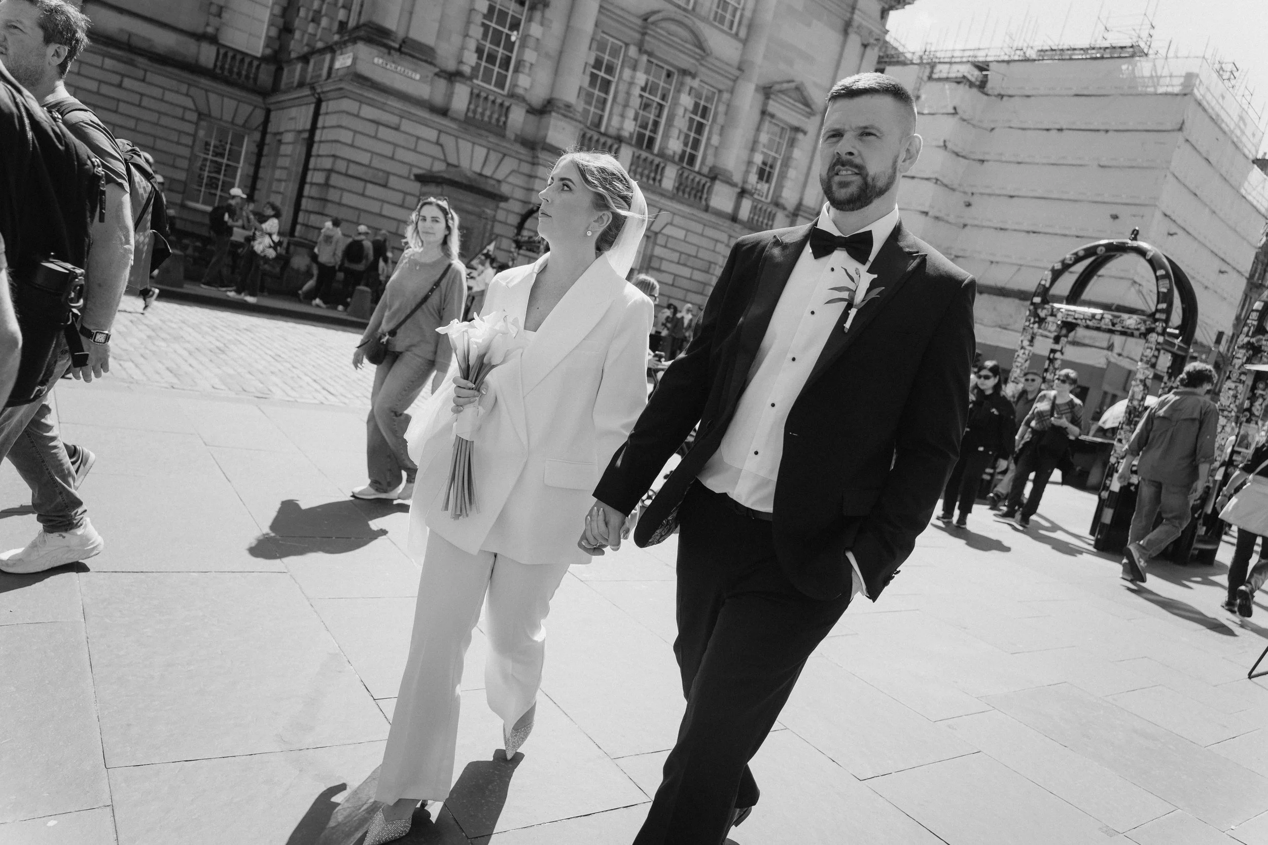 A wedding couple, a man in a tuxedo and a woman in a white suit, walking hand in hand outdoors in a city street, with onlookers around. - captured by an Edinburgh wedding photographer