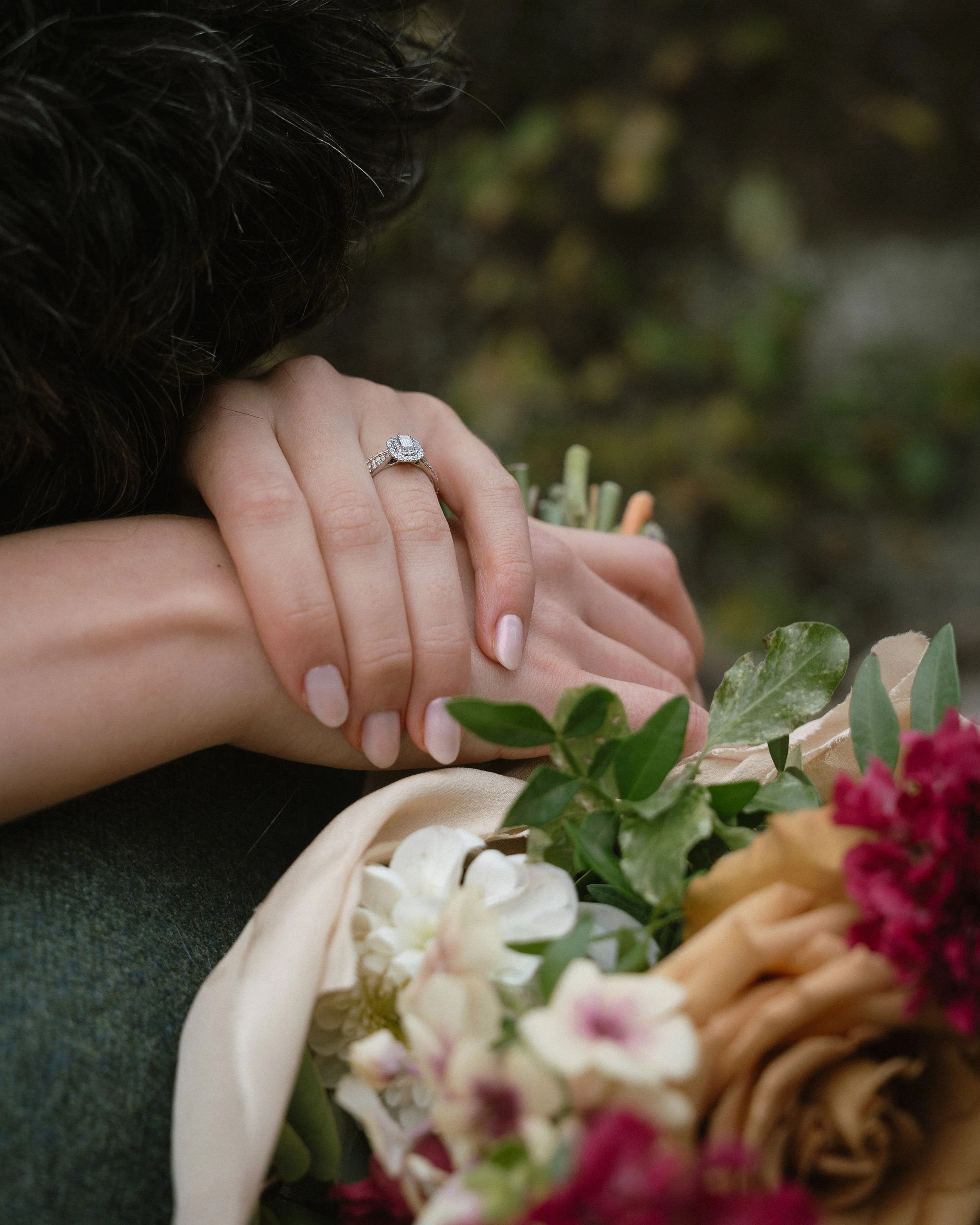 Close-up of a woman with wavy dark hair resting her hand on top of another person's hand, both hands on a bouquet of flowers, with an engagement ring on her finger, outdoors.