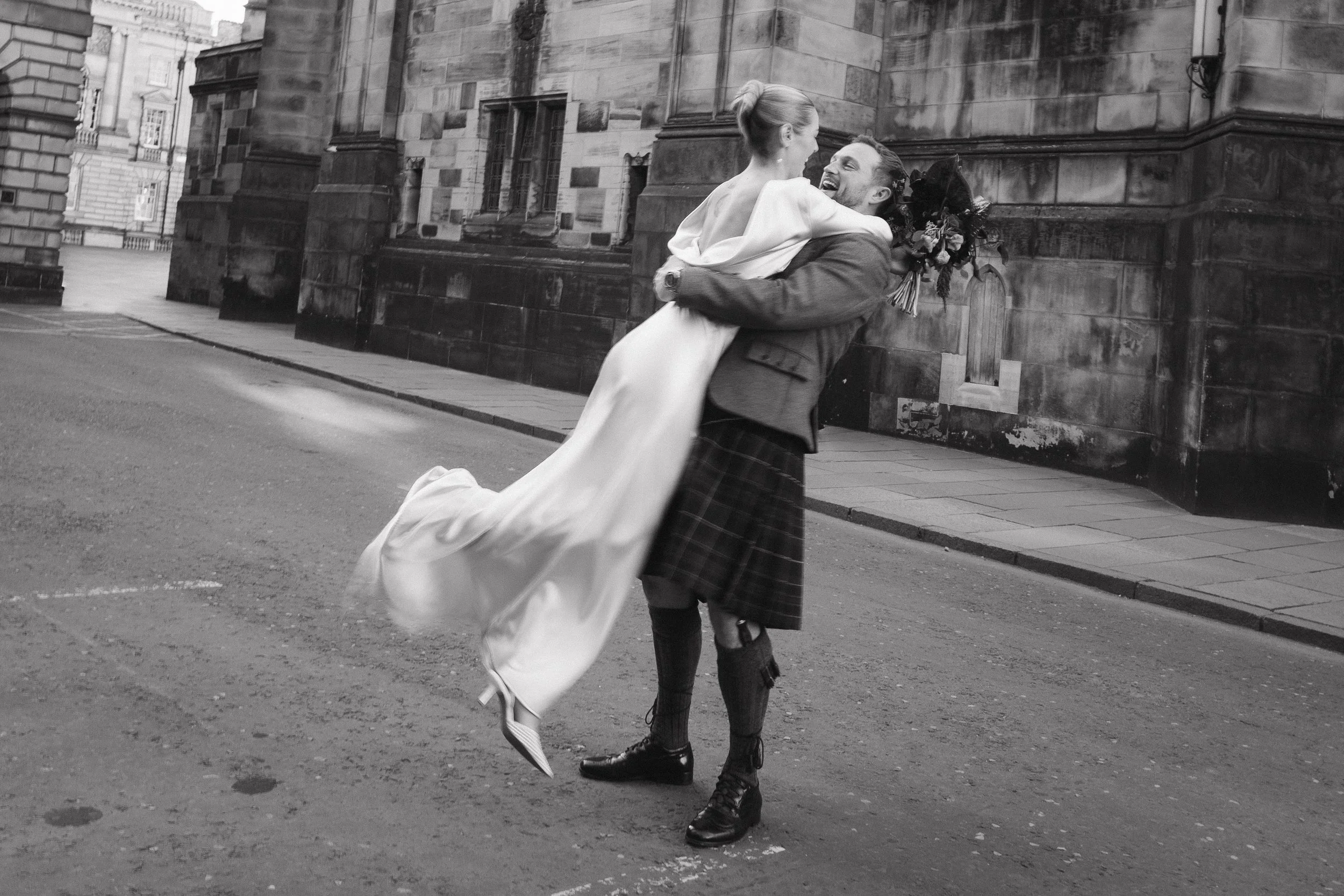 A man in traditional Scottish attire, wearing a kilt, lifts and dances with a woman in a long wedding dress on a city street, both smiling. - captured by an Edinburgh wedding photographer
