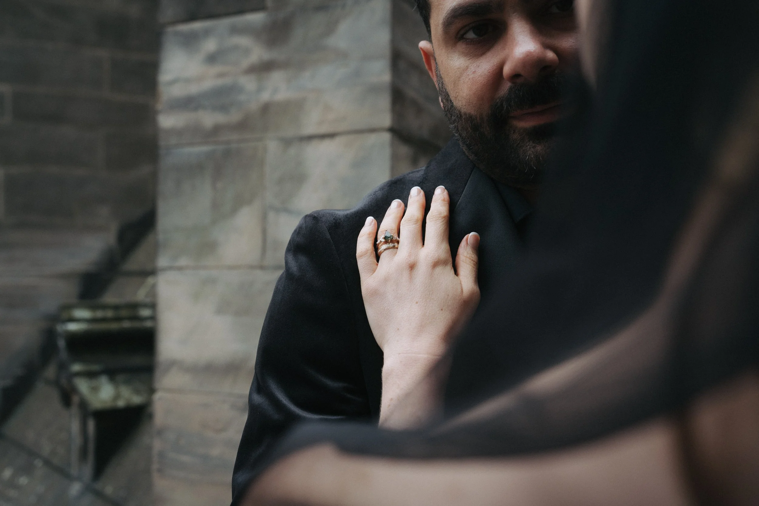 A woman with a wedding ring placing her hand on a man's shoulder. The man has a beard and is wearing a black suit, and they are indoors with a stone wall background. - captured by an Edinburgh wedding photographer