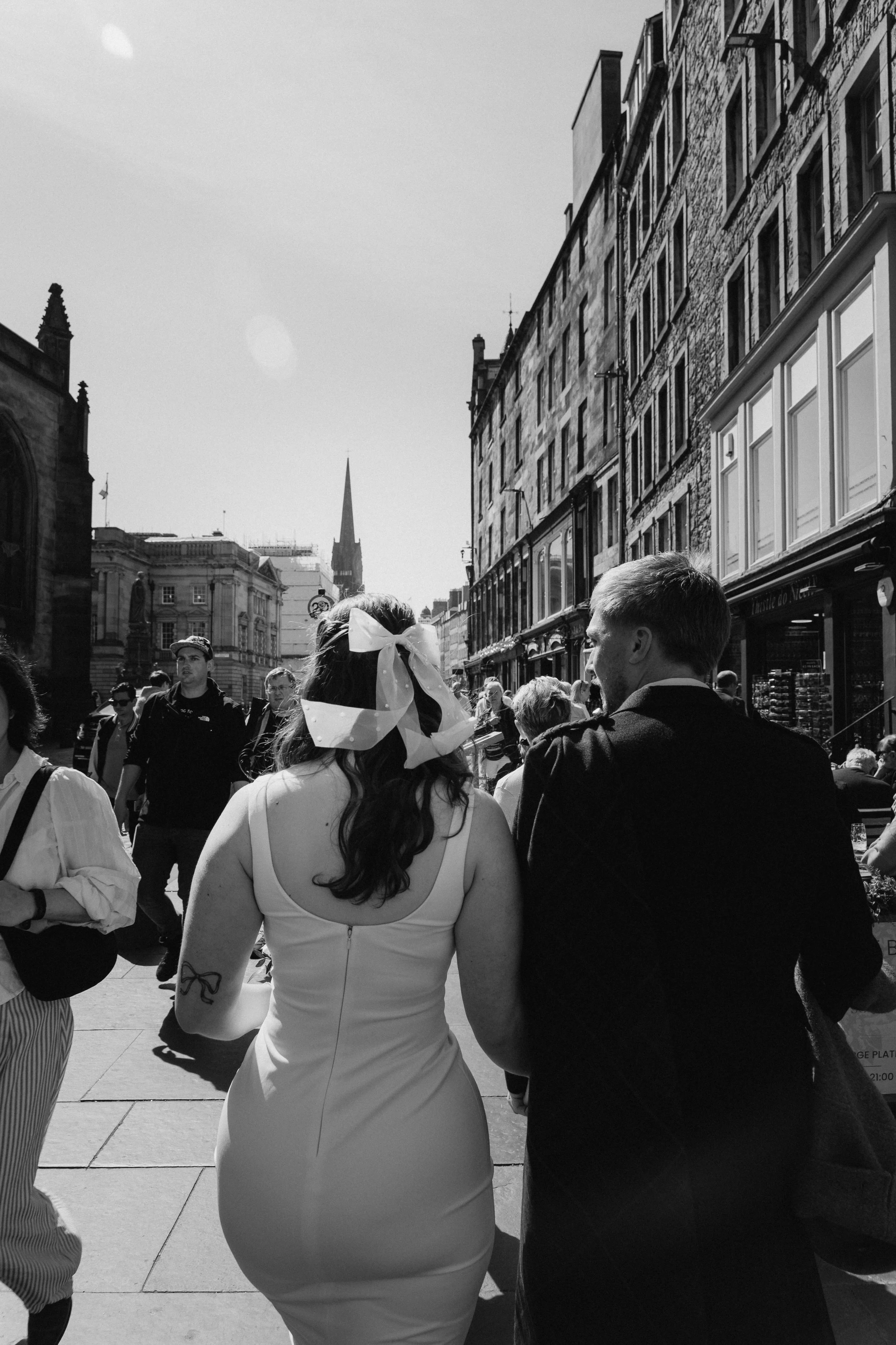 A black and white photo of a woman and a man walking on a busy city street, seen from behind, with the woman wearing a dress and a large bow in her hair, and the man in a suit. - captured by an Edinburgh wedding photographer