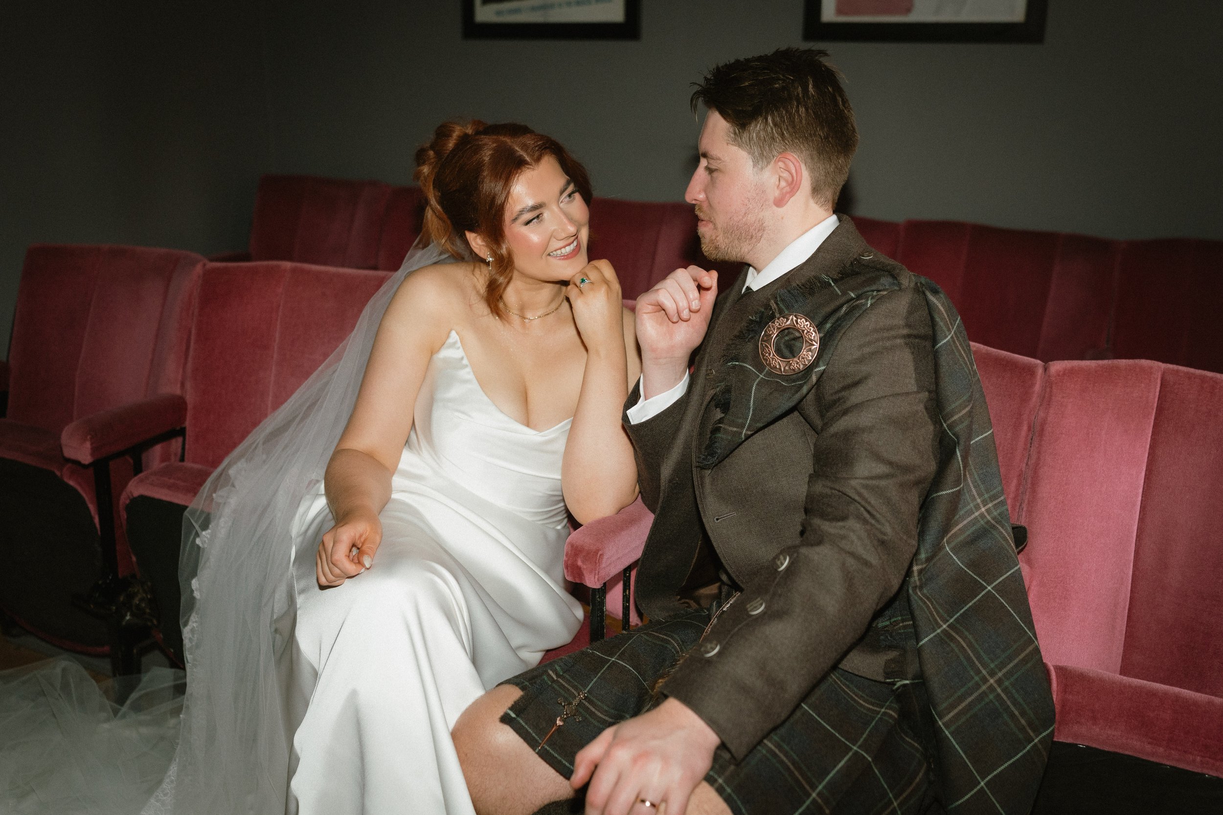 A bride and groom sitting close together on red velvet theater seats, smiling and gazing at each other in a dimly lit theater. - captured by an Edinburgh wedding photographer