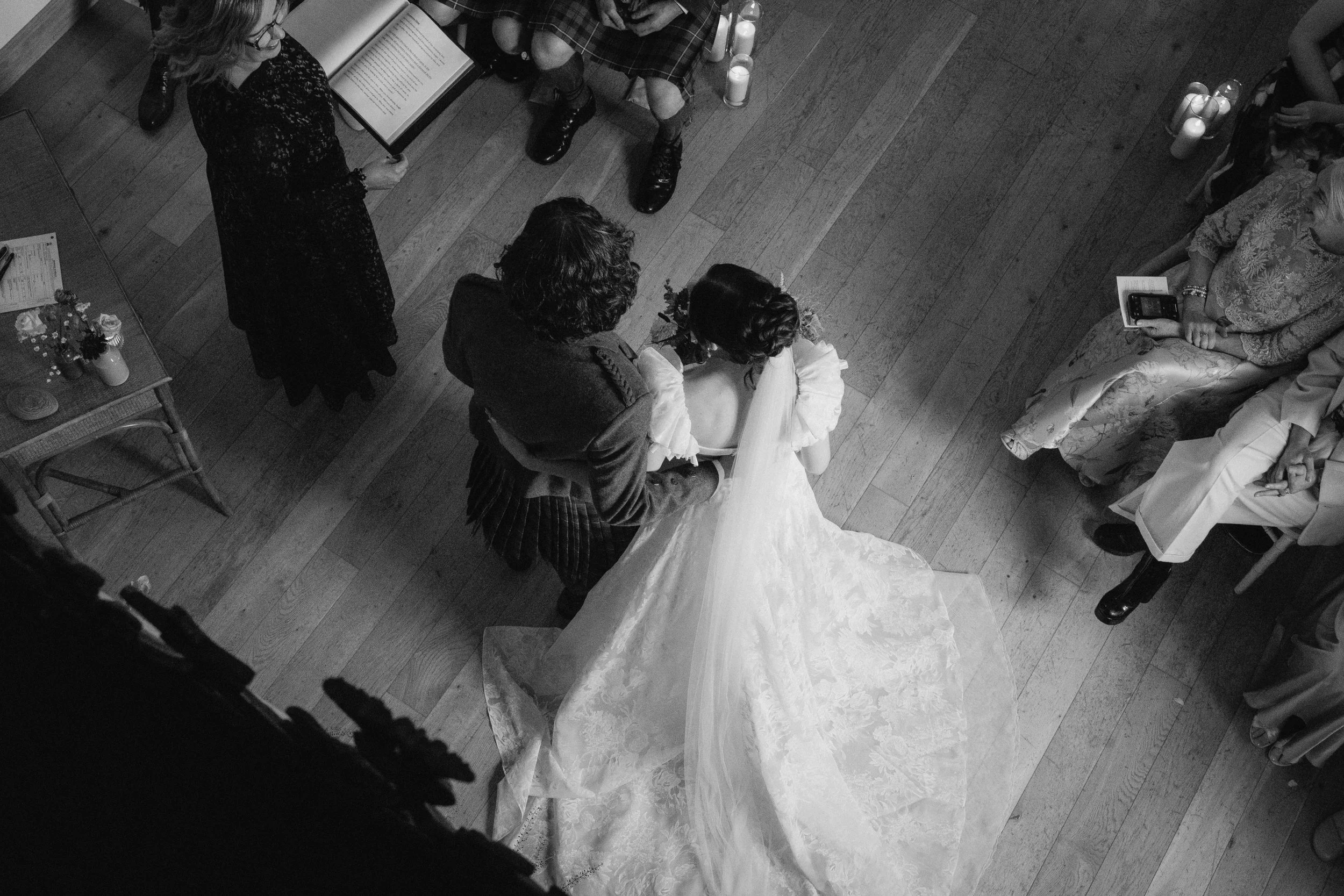 Black and white photo of a wedding ceremony from above, with the bride in a wedding gown embracing a person, likely the groom, while a woman in a traditional dress and other guests observe. - captured by an Edinburgh wedding photographer