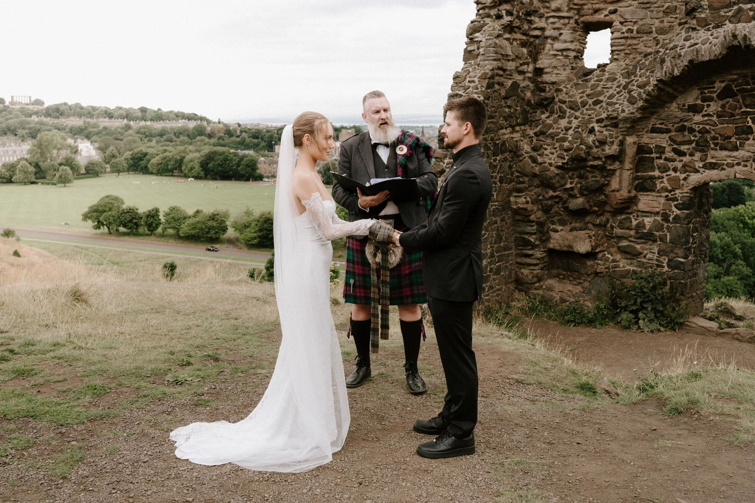 A wedding ceremony outdoors with a bride and groom holding hands and facing each other, an officiant in traditional Scottish attire, and a scenic landscape with greenery and a stone ruin in the background. - captured by an Edinburgh wedding photograp