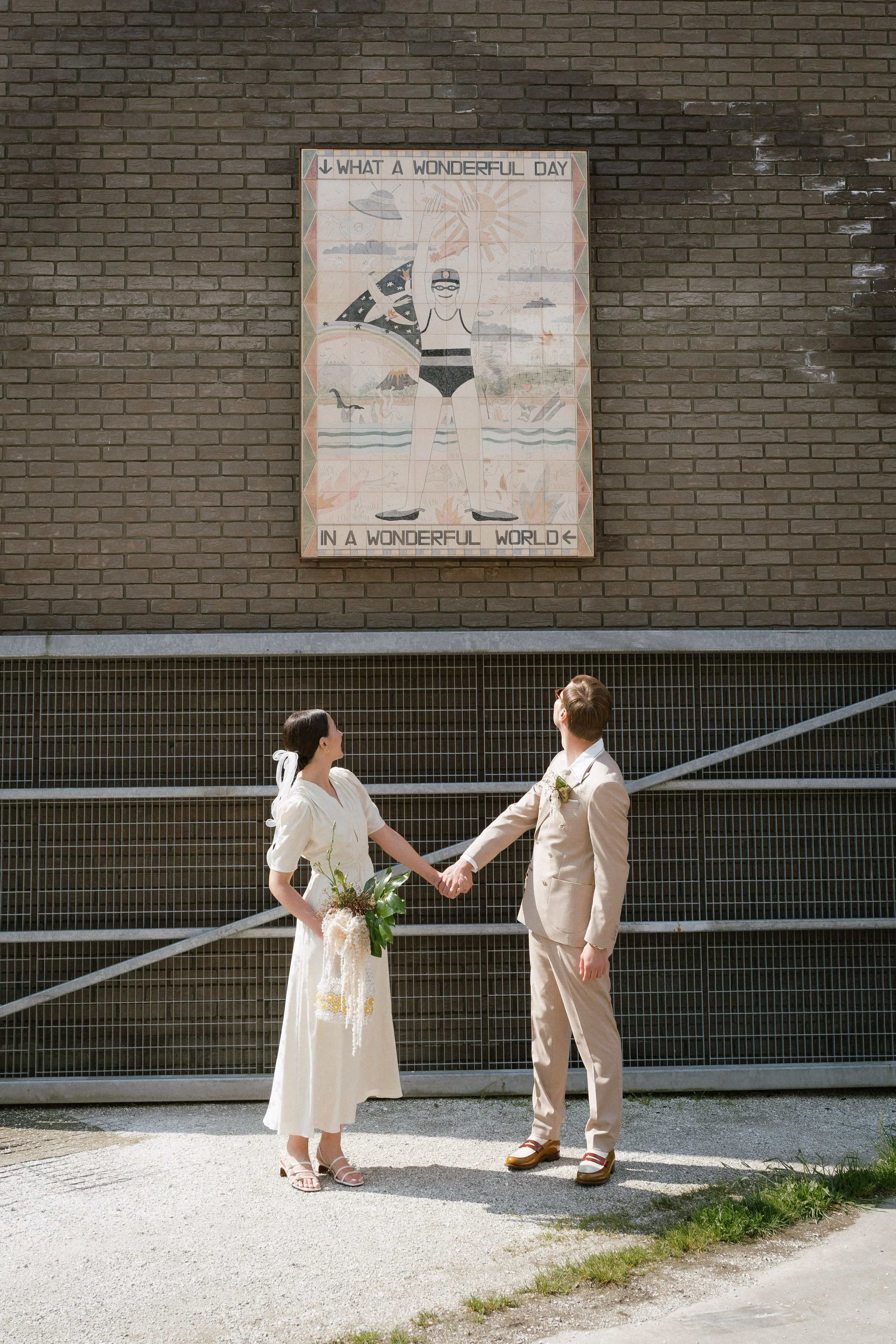 A bride and groom holding hands and looking at a mural on a brick wall. The mural shows a person in a swimsuit at the beach with text saying "What a wonderful day in a wonderful world."  - captured by an Edinburgh wedding photographer