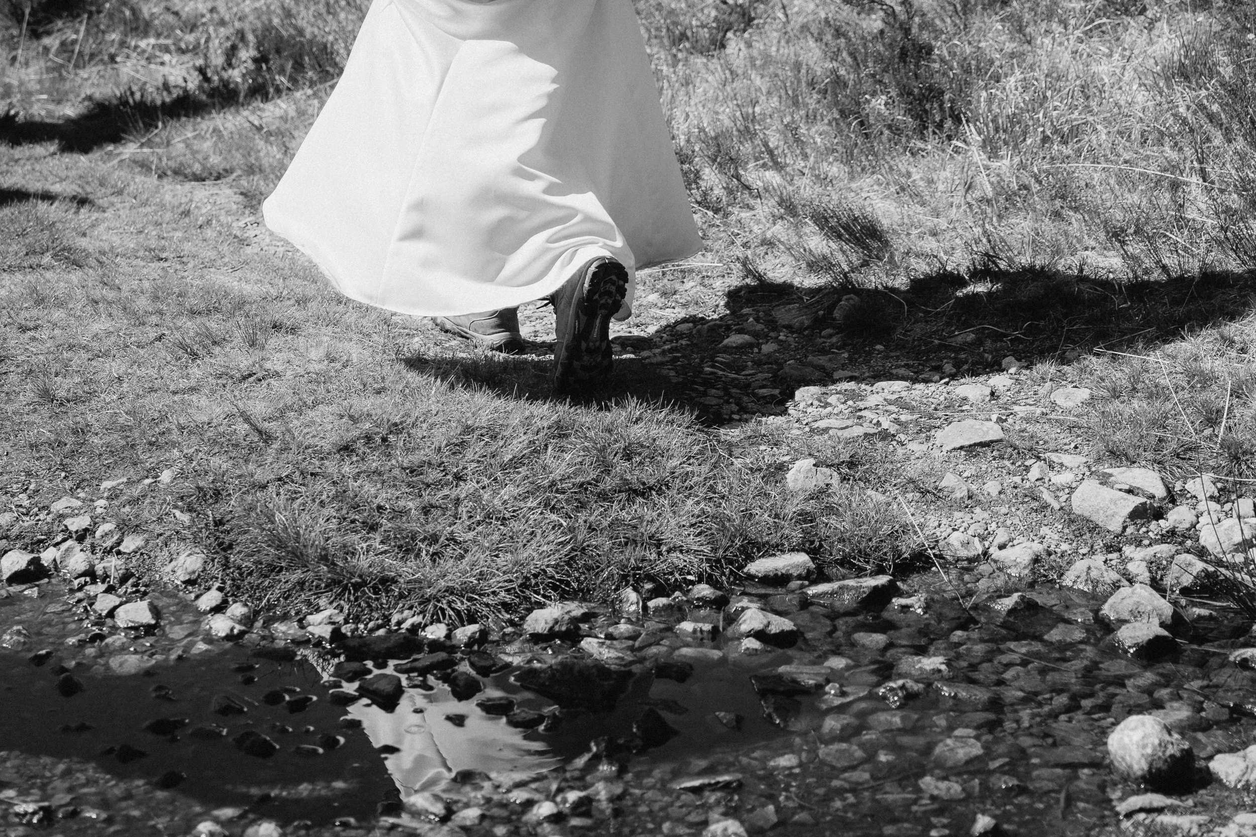 Person walking on rocky terrain near a small stream, wearing a flowing white dress and hiking boots, casting a shadow on the ground. - captured by an Edinburgh wedding photographer