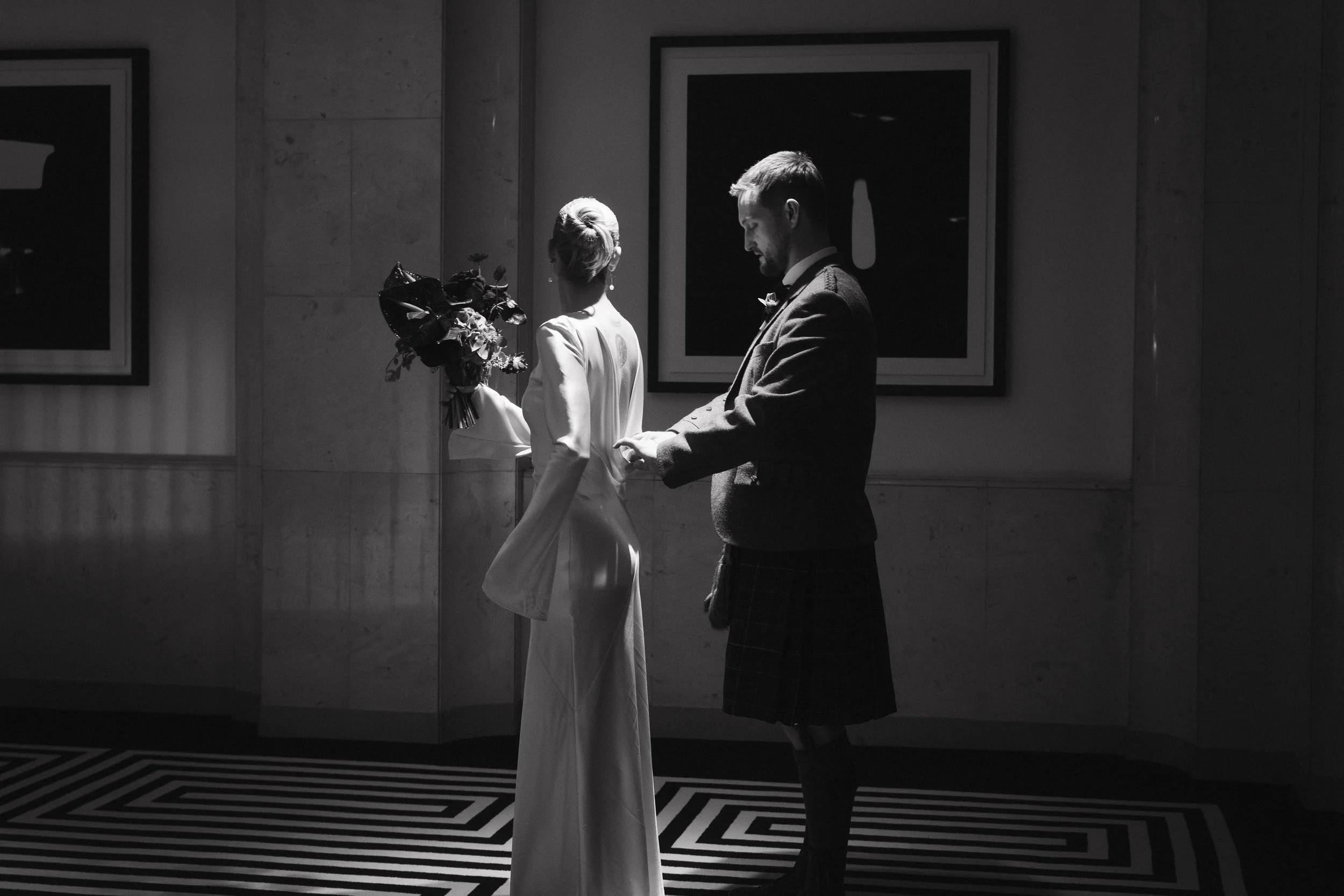 A black and white photo of a woman in a white dress holding a bouquet of flowers and a man in a plaid kilt and jacket standing in a hallway with framed artwork on the wall. - captured by an Edinburgh wedding photographer