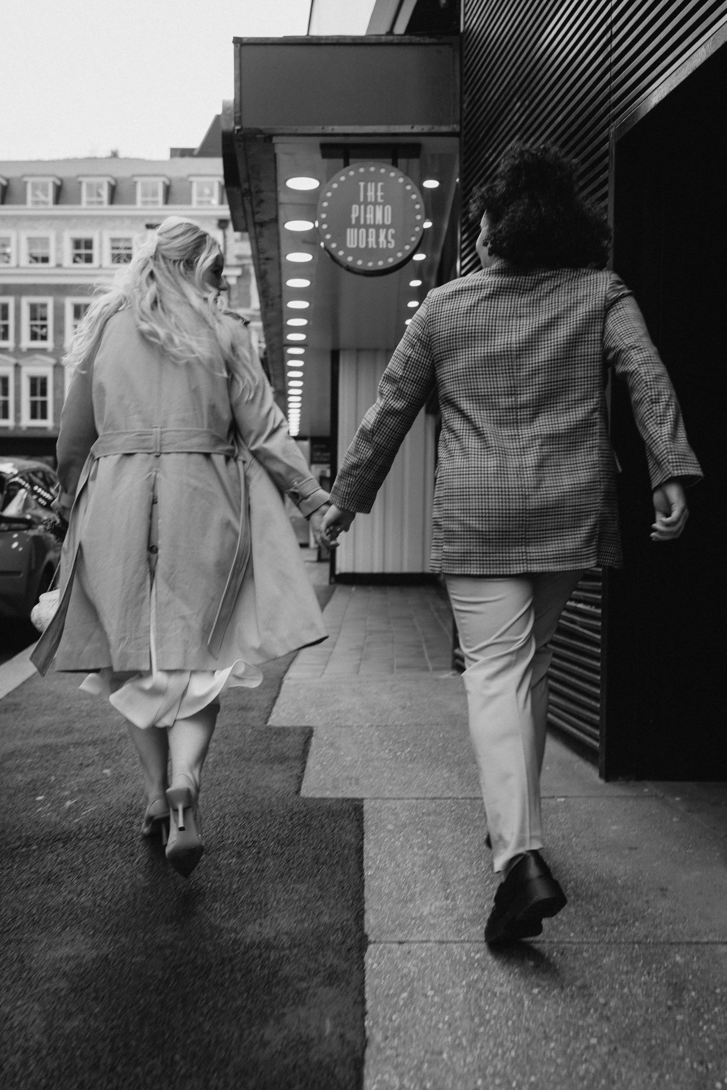 Two women walking hand in hand down a city sidewalk, one with long hair wearing a trench coat and high heels, the other with curly hair wearing a checkered blazer and light pants, near a sign that reads 'The Piano Works'.