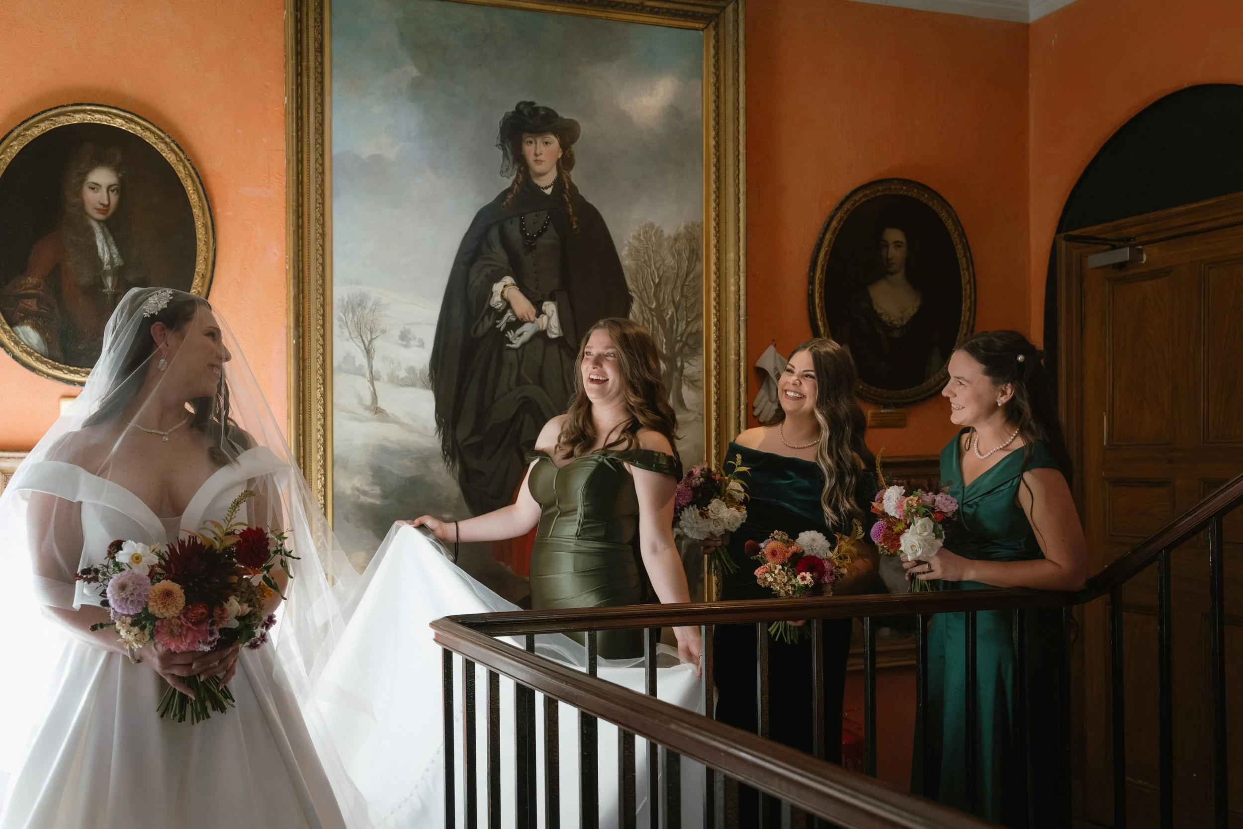 A bride in a white wedding gown holding a bouquet of flowers stands with four bridesmaids inside an elegant building with framed portraits on the walls. The bridesmaids wear dark green and black dresses and hold bouquets, smiling and laughing with th