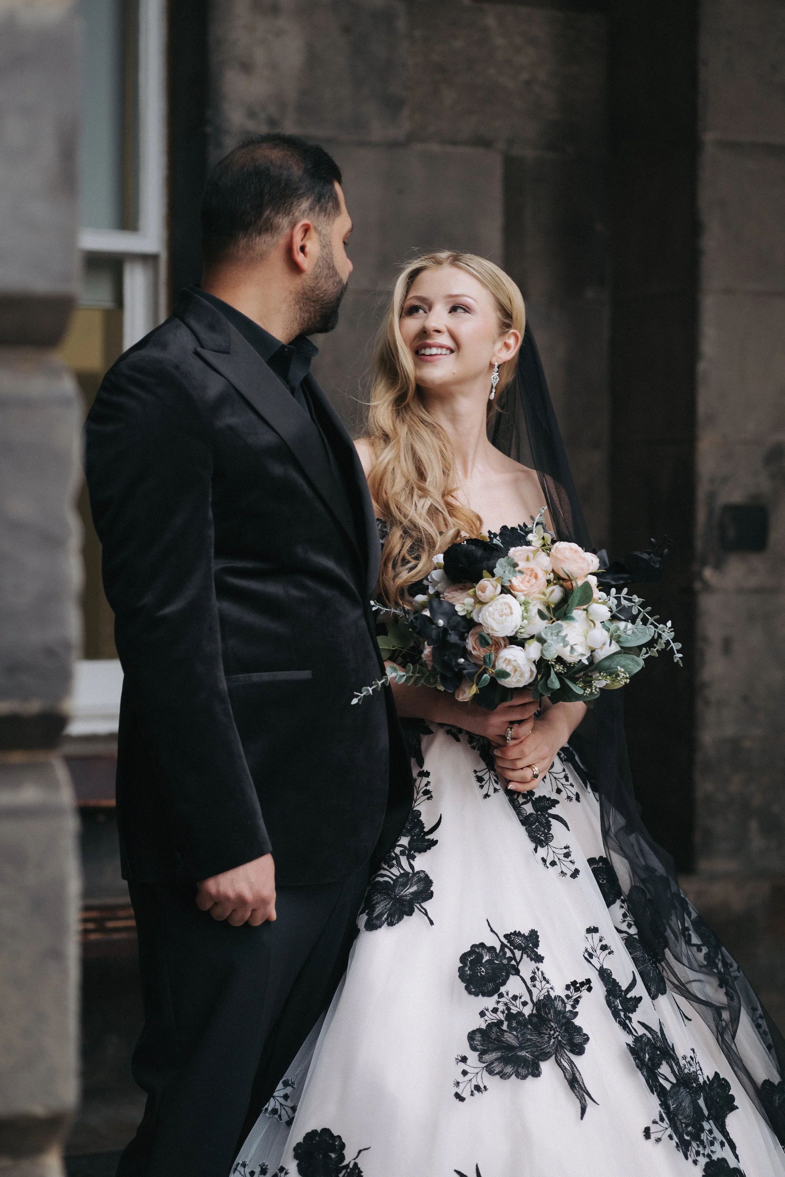 A bride and groom on their wedding day, talking and smiling at each other outside, with stone walls around them. The bride is holding a bouquet of white and pink flowers and wearing a white gown with black floral embroidery, while the groom is in a b