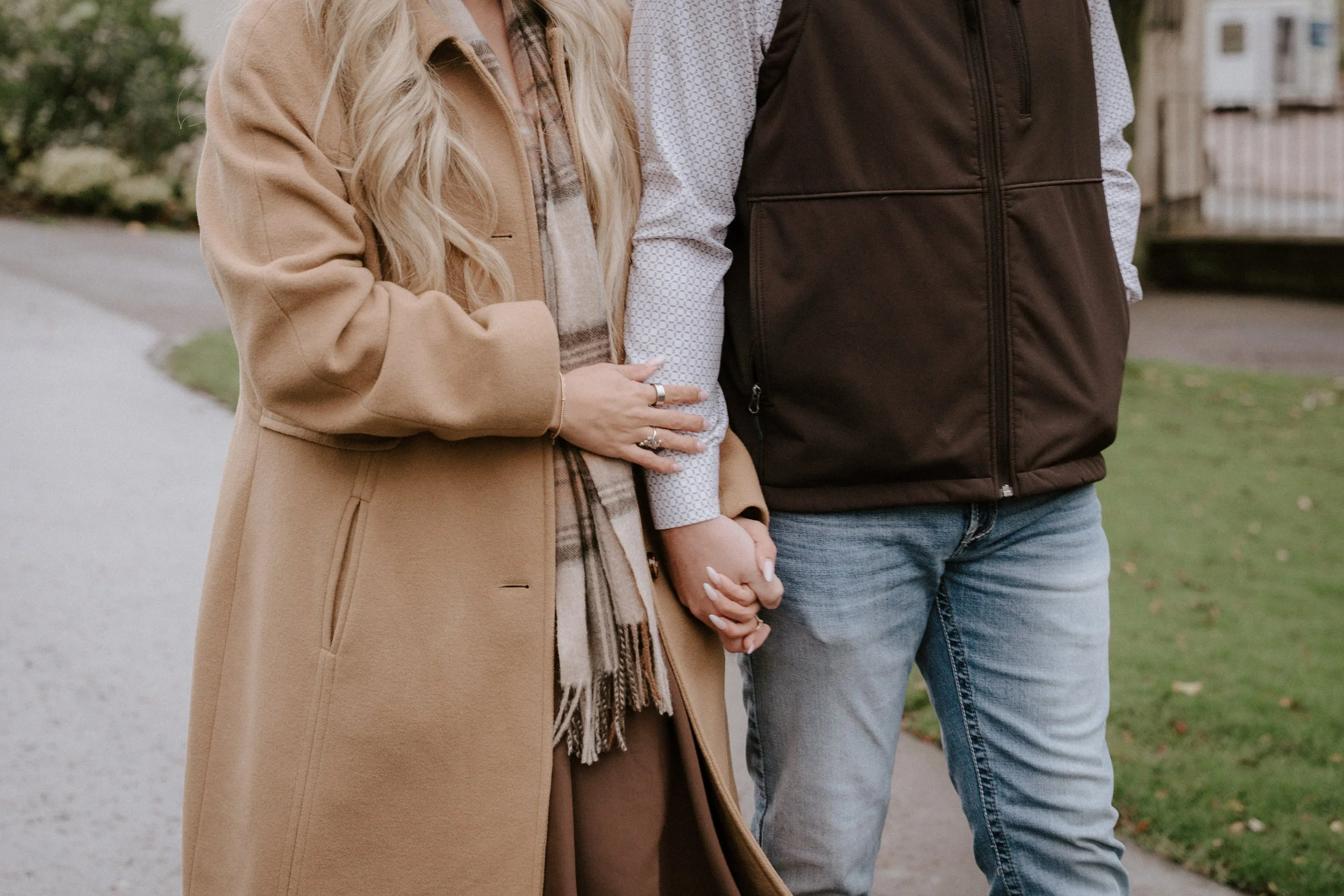 A couple holding hands standing on a sidewalk outdoors, woman wearing a beige coat and plaid scarf, man wearing a dark vest and jeans, visible in a park or residential area - captured by an Edinburgh wedding photographer