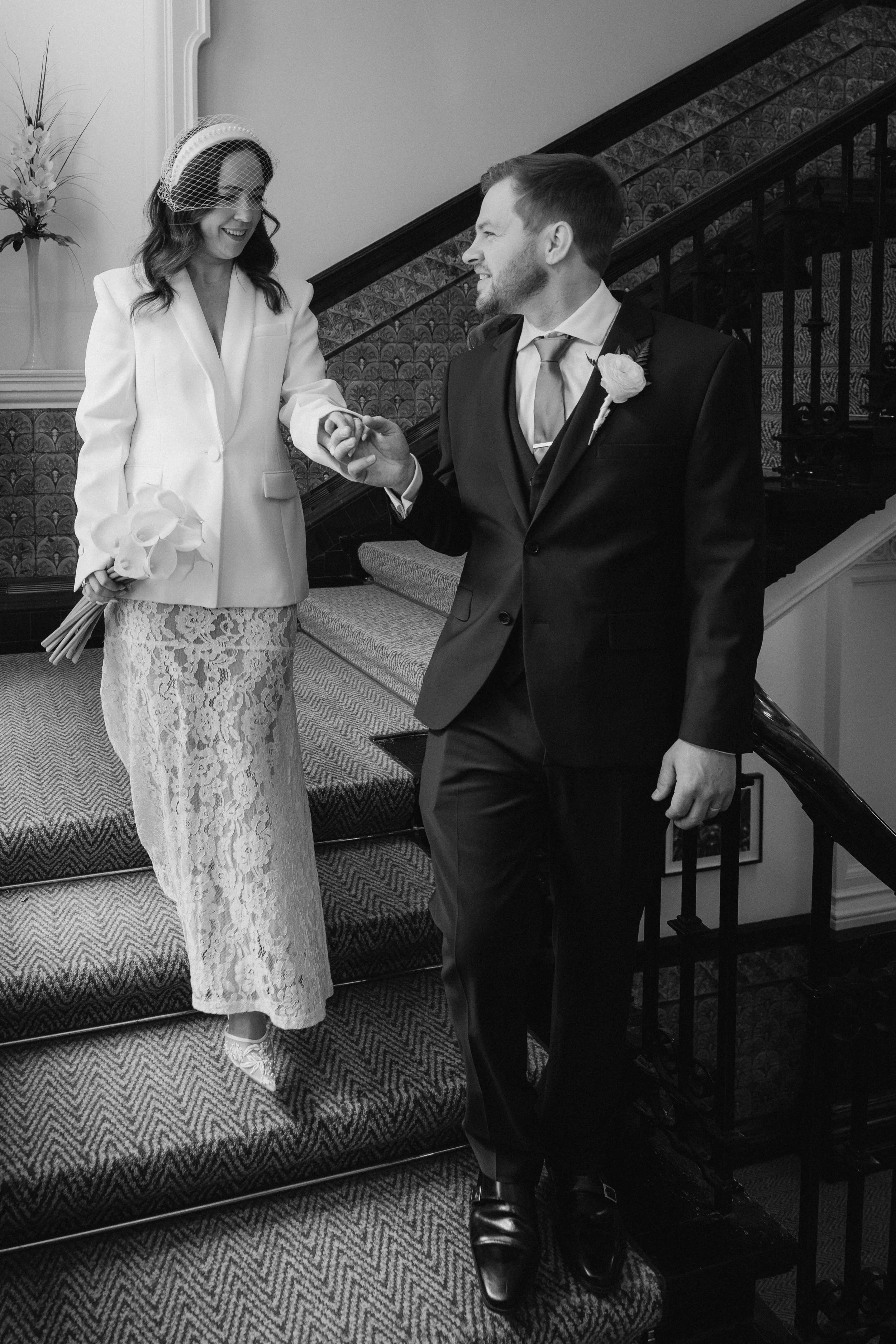 Black and white photo of a bride in a lace dress and blazer holding a bouquet, smiling as she holds hands with a groom in a tuxedo with a boutonniere, descending stairs inside a decorated interior. - captured by an Edinburgh wedding photographer
