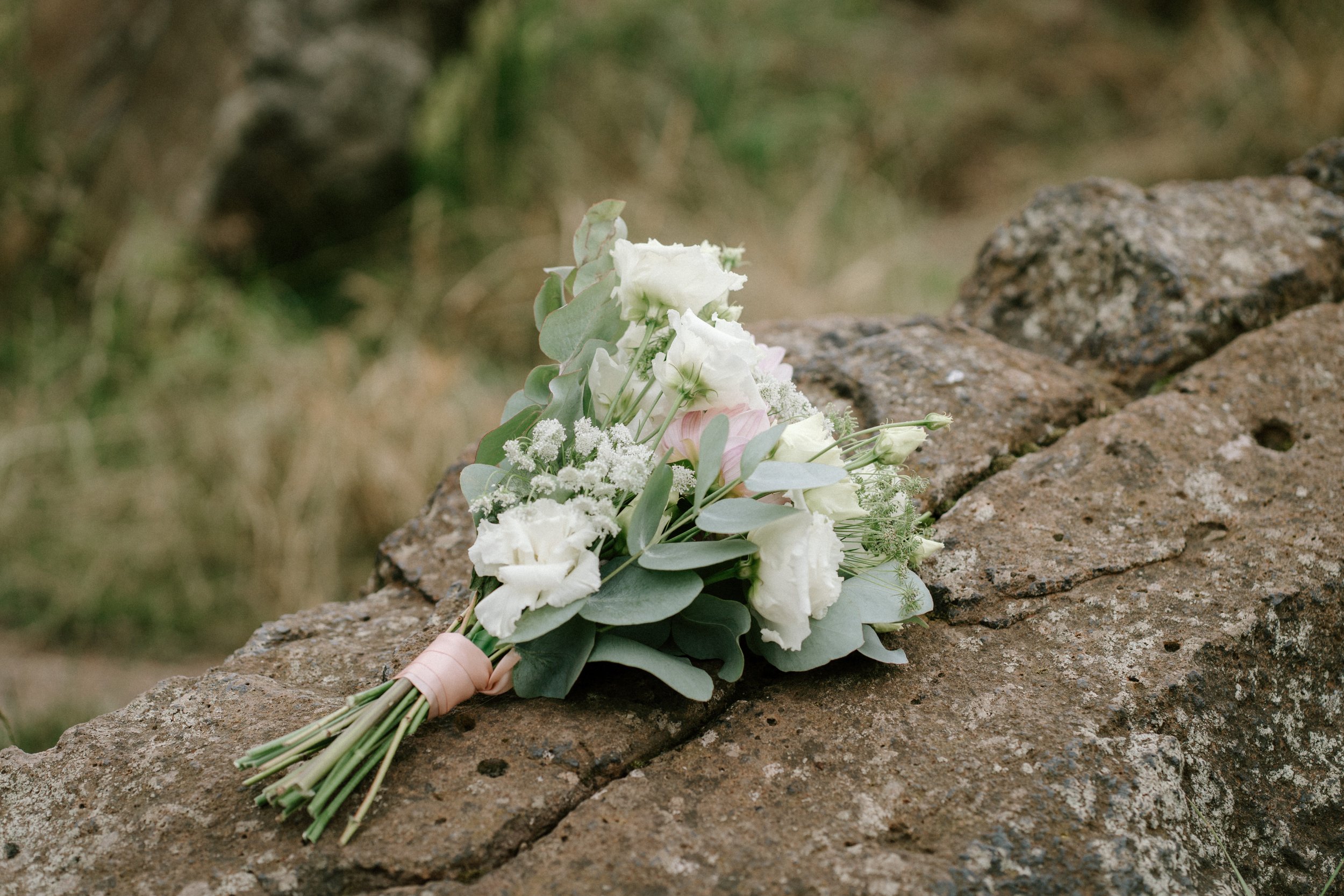 A bridal bouquet with white flowers and green leaves, wrapped with a pink ribbon, resting on a stone surface outdoors. - captured by an Edinburgh wedding photographer