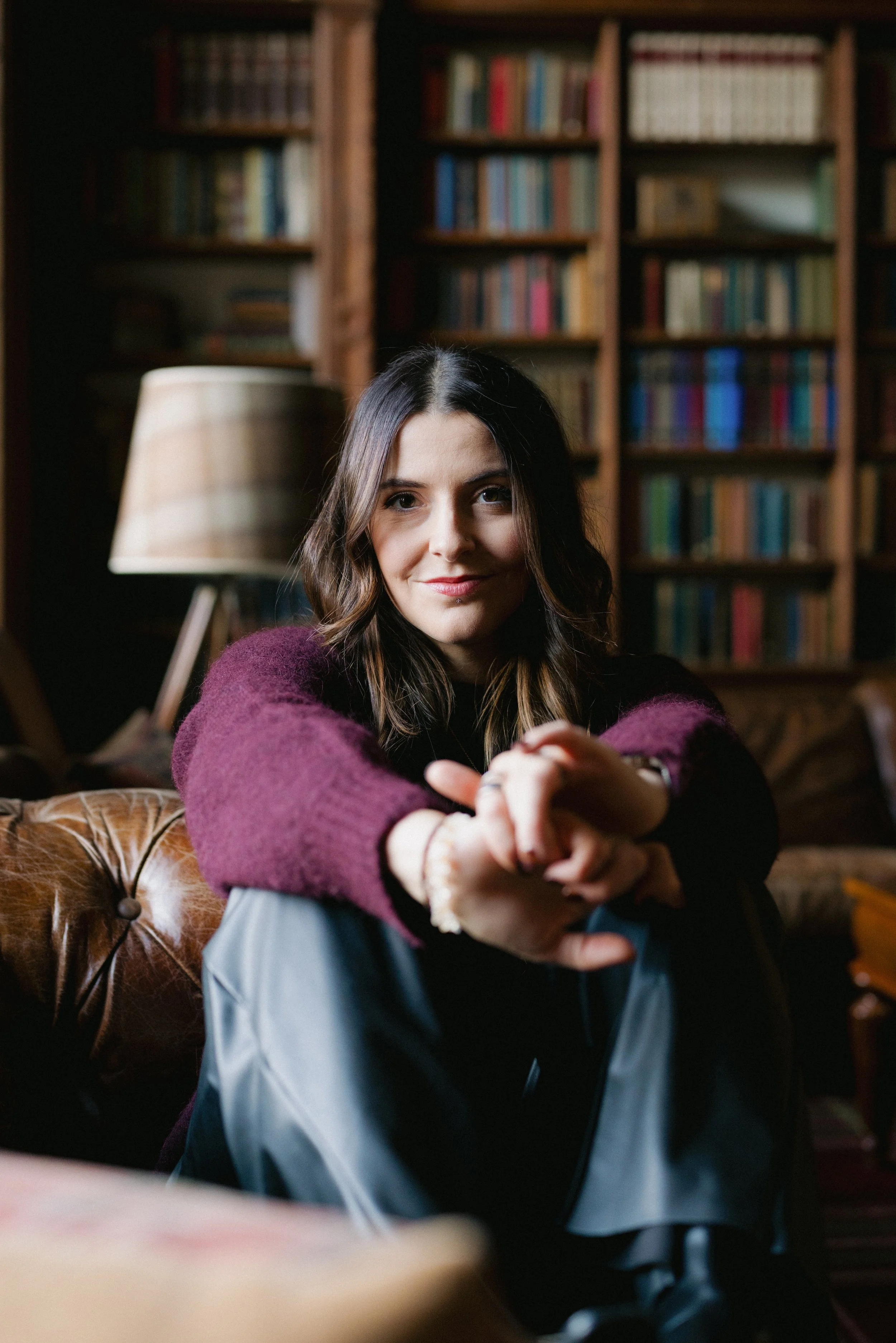 A woman with dark wavy hair sitting on a leather sofa in a cozy room with wooden bookshelves filled with books in the background. She is wearing a maroon sweater and smiling at the camera.