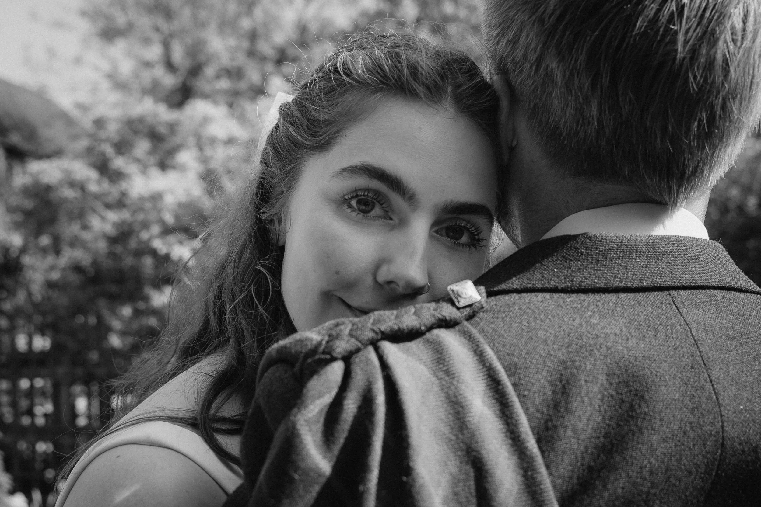 A woman with wavy hair and a septum piercing smiling while resting her head on a man's shoulder, in an outdoor setting with trees in the background. - captured by an Edinburgh wedding photographer