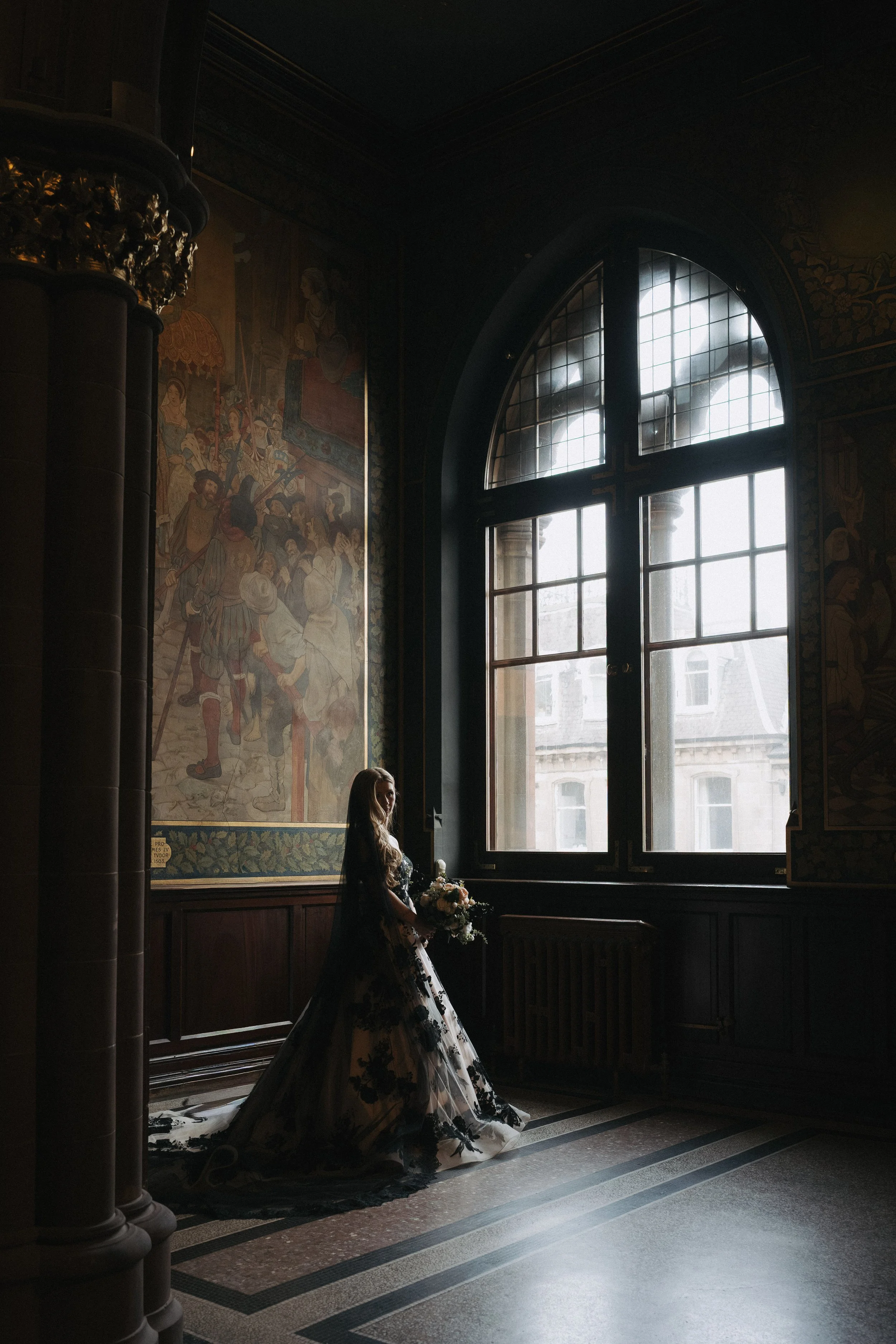 A woman in a black floral wedding dress holding a bouquet of flowers stands inside a dim, ornate room with a large arched window, historic murals on the wall, and dark wood paneling. - captured by an Edinburgh wedding photographer
