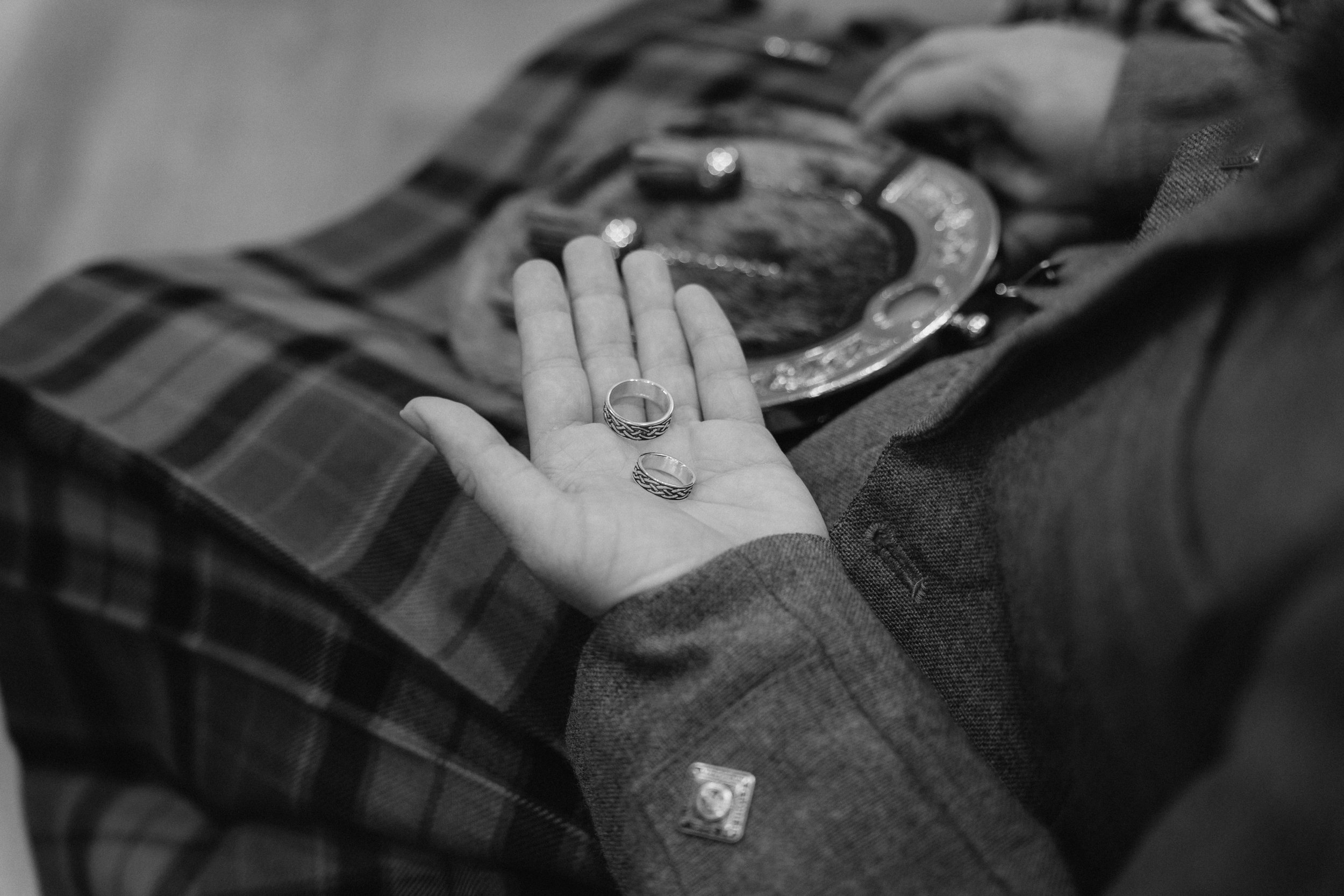 A person dressed in a plaid shirt and blazer, lying down with a sheriff's badge on chest, holding two rings on their open palm, with a sheriff's badge and a hat visible. - captured by an Edinburgh wedding photographer