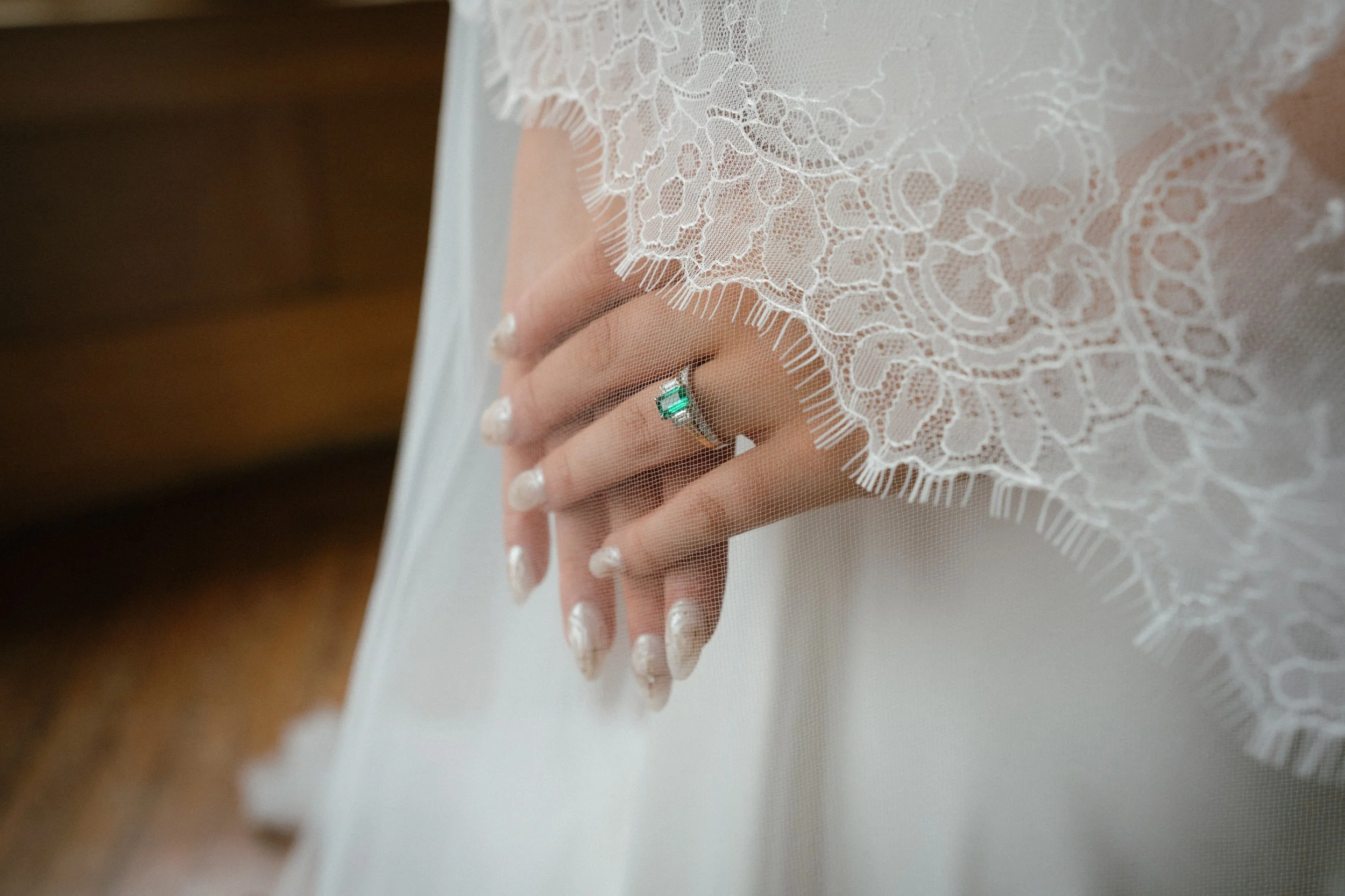 Close-up of a hand with a ring that has a green gemstone, partially covered by a delicate lace fabric of a wedding dress. - captured by an Edinburgh wedding photographer