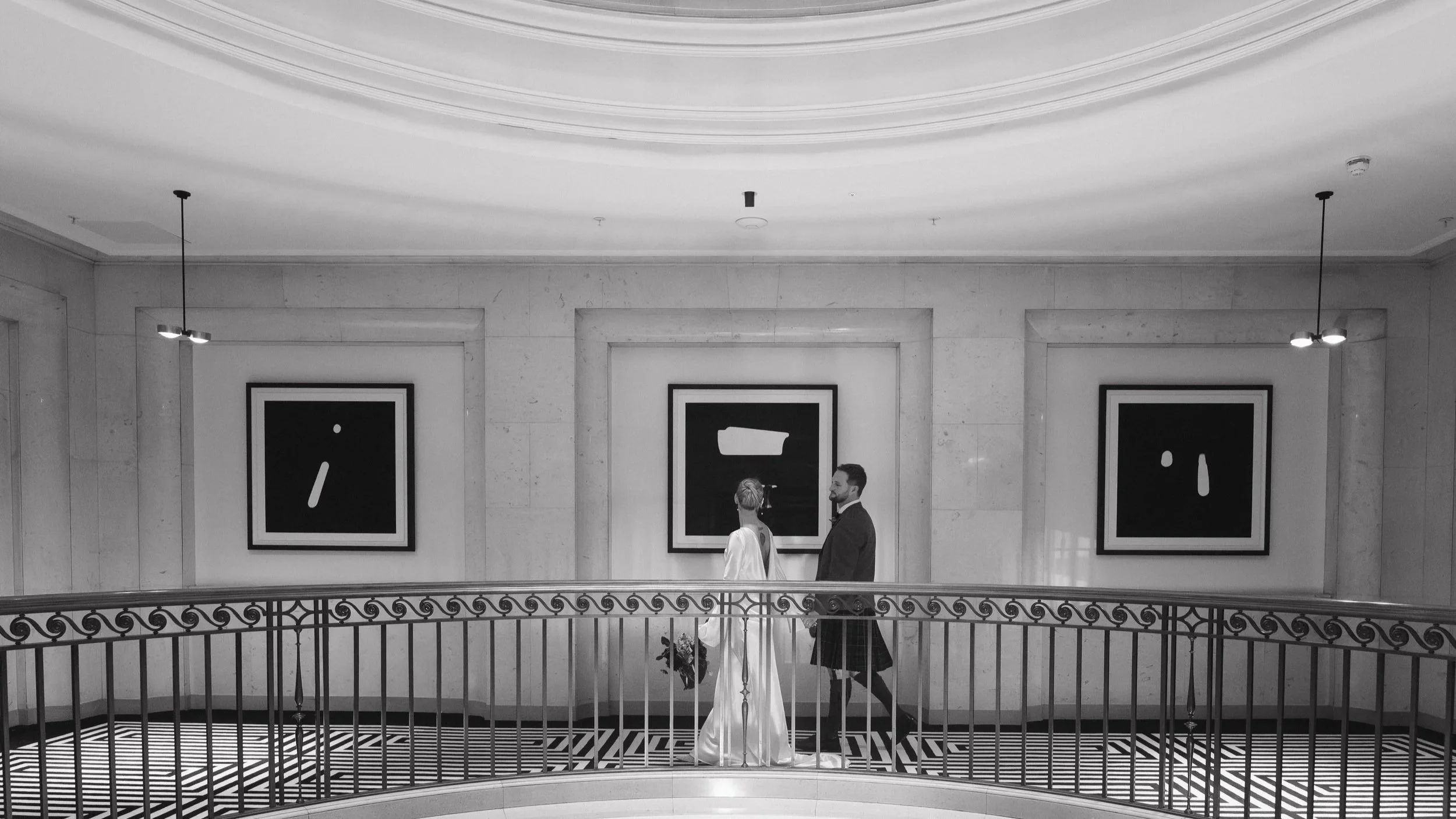 A couple in wedding attire walking in an art gallery or museum with black and white abstract paintings hanging on the wall behind them - captured by an Edinburgh wedding photographer