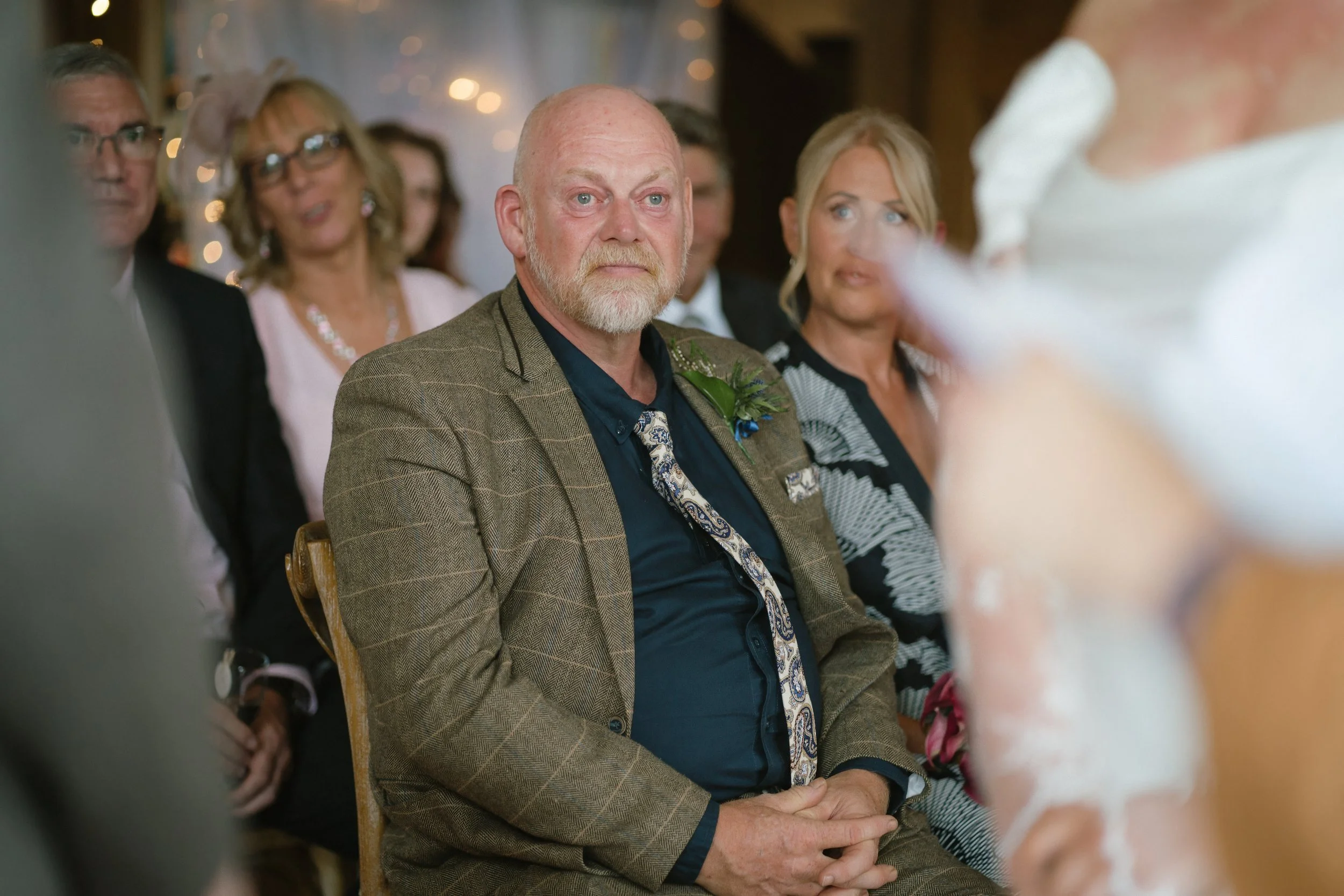 A middle-aged man with a beard and bald head, wearing a brown plaid suit and a patterned tie, sitting at a wedding or formal event, looking serious. Behind him are other guests, including a woman with blonde hair and a black and white dress, and a ma