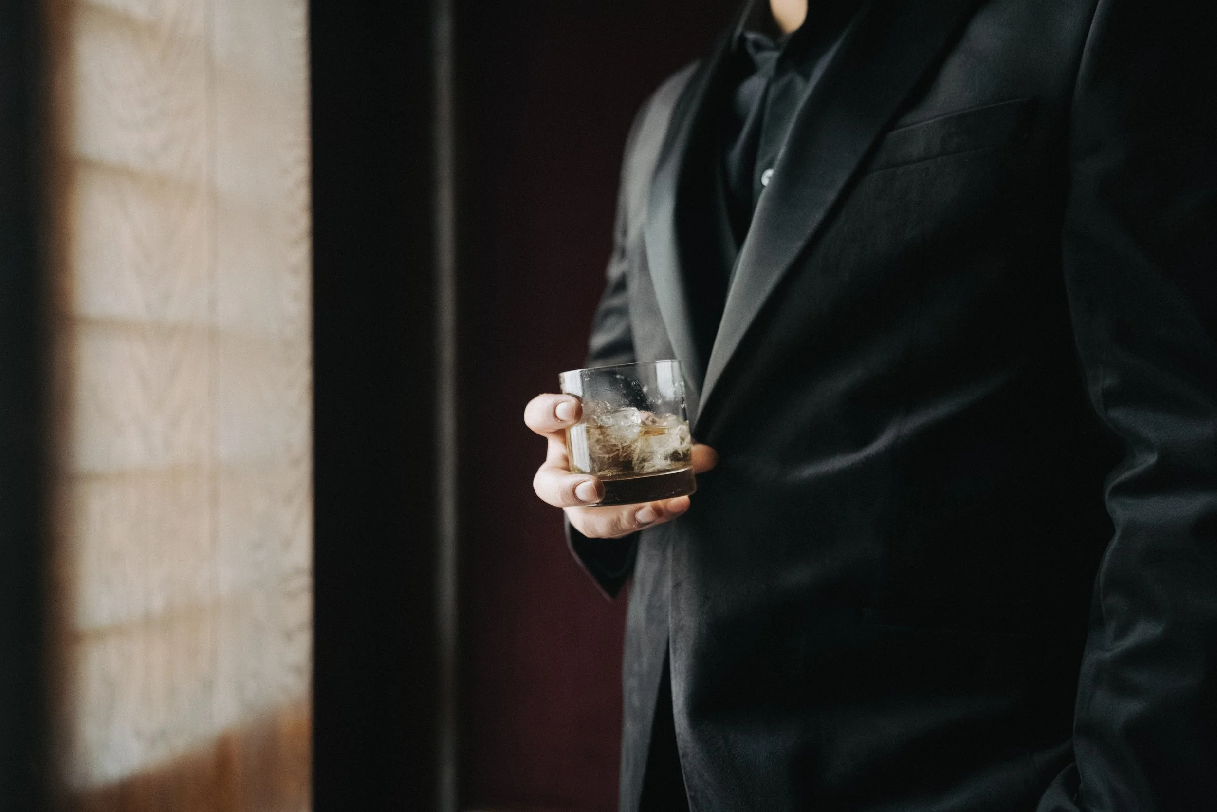 A person wearing a black suit holding a glass of dark liquor with ice, standing against a dark background. - captured by an Edinburgh wedding photographer