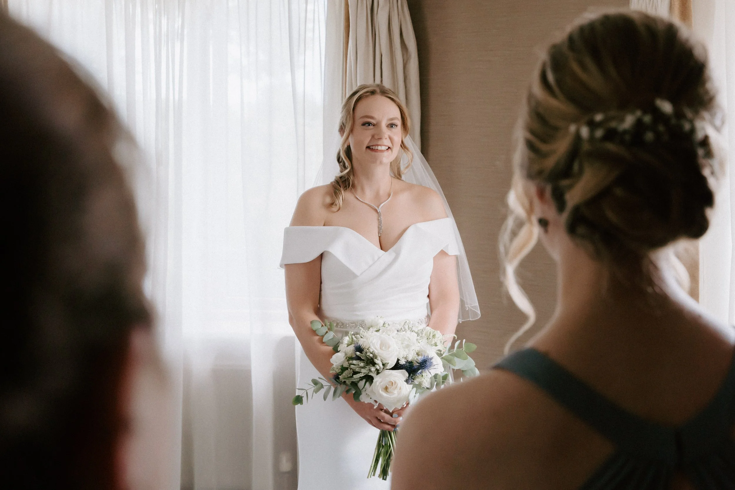 A bride in a white off-shoulder wedding dress holding a bouquet of white roses and greenery, smiling in front of sheer curtains while two women, one with a dark dress and the other with a braided hairstyle, look at her.