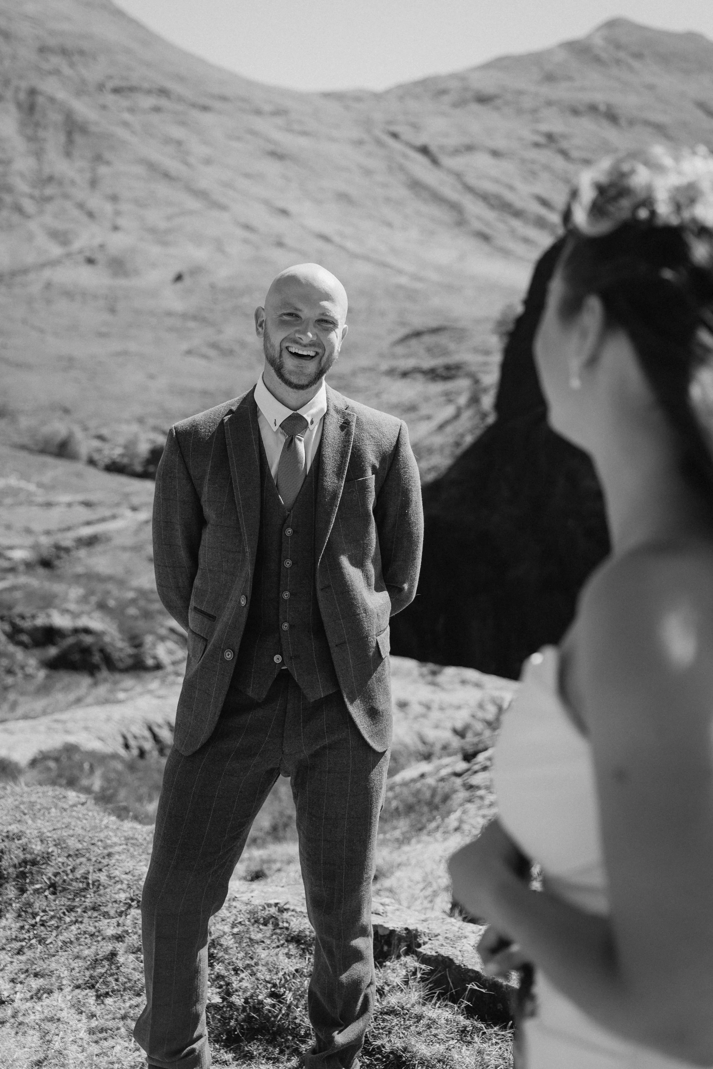 A smiling man in a suit stands outdoors in a mountainous landscape with a woman with dark hair in the foreground. - captured by an Edinburgh wedding photographer