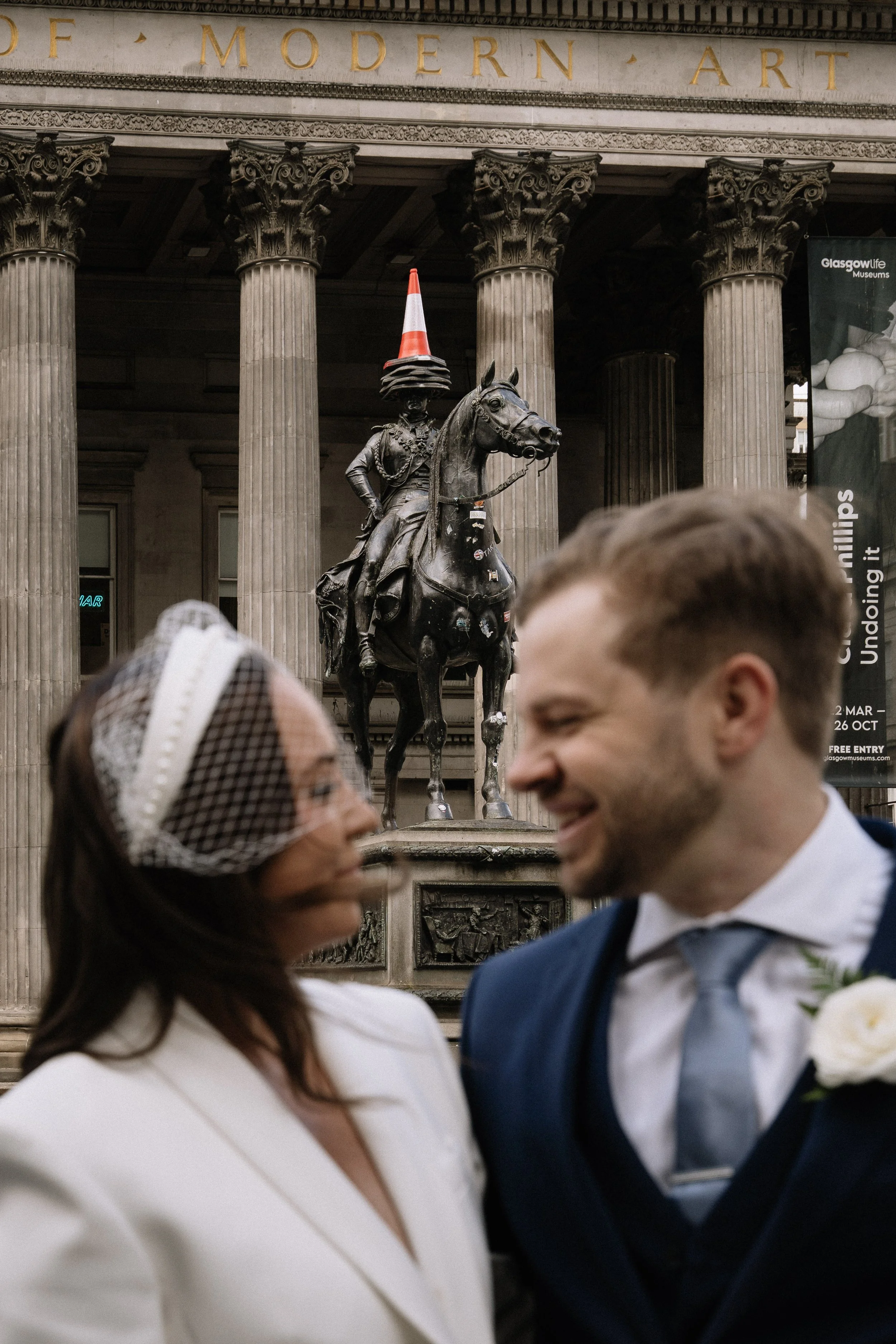 A couple dressed for a wedding or formal event standing in front of a statue of a man on horseback at the Glasgow Art Museum, with columns and a sign about modern art in the background.