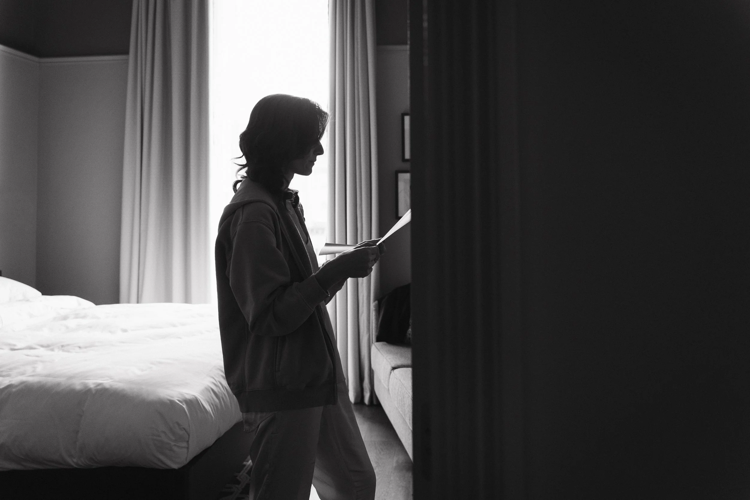 A person standing in a bedroom, reading a letter or paper, with a bed and window with curtains in the background. - captured by an Edinburgh wedding photographer
