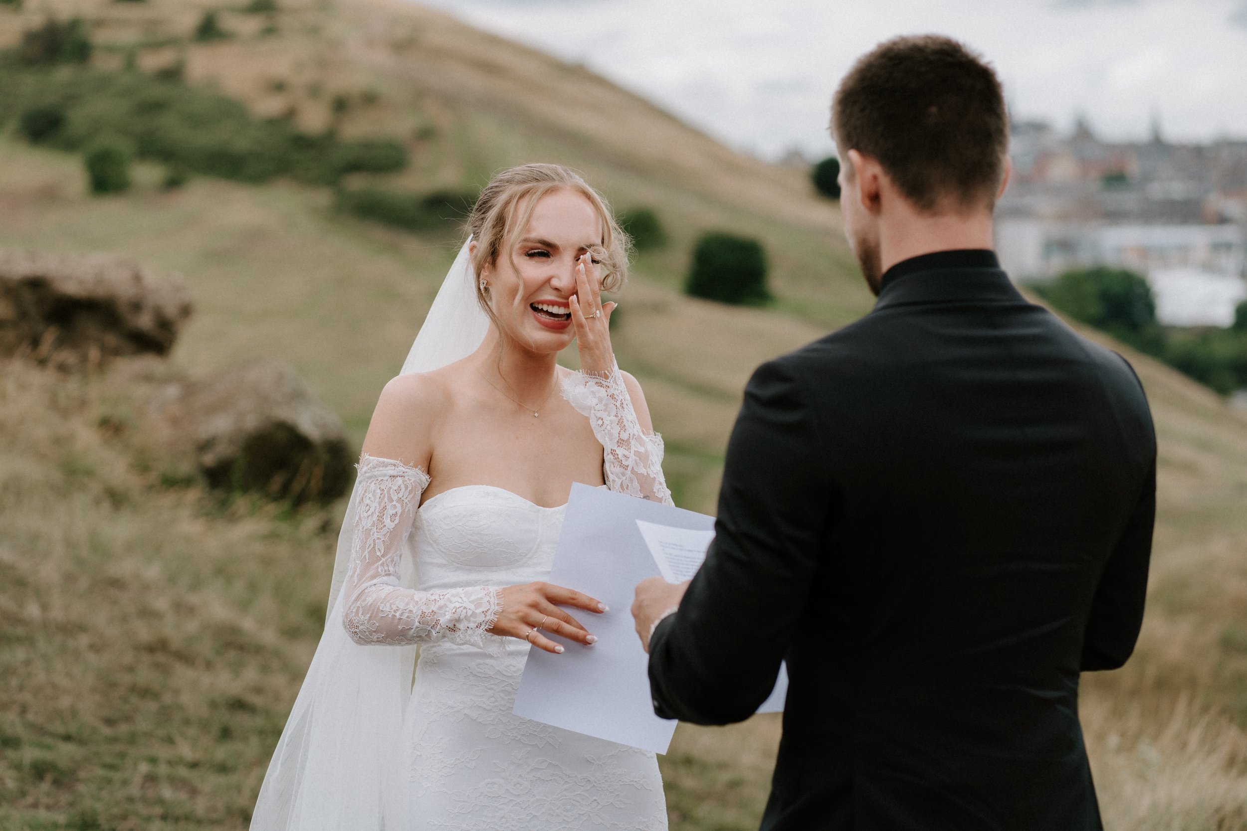 A bride in a white wedding dress with lace sleeves is crying tears of joy while holding a piece of paper, standing outdoors on a hillside, during her wedding vows with a groom in a black suit.