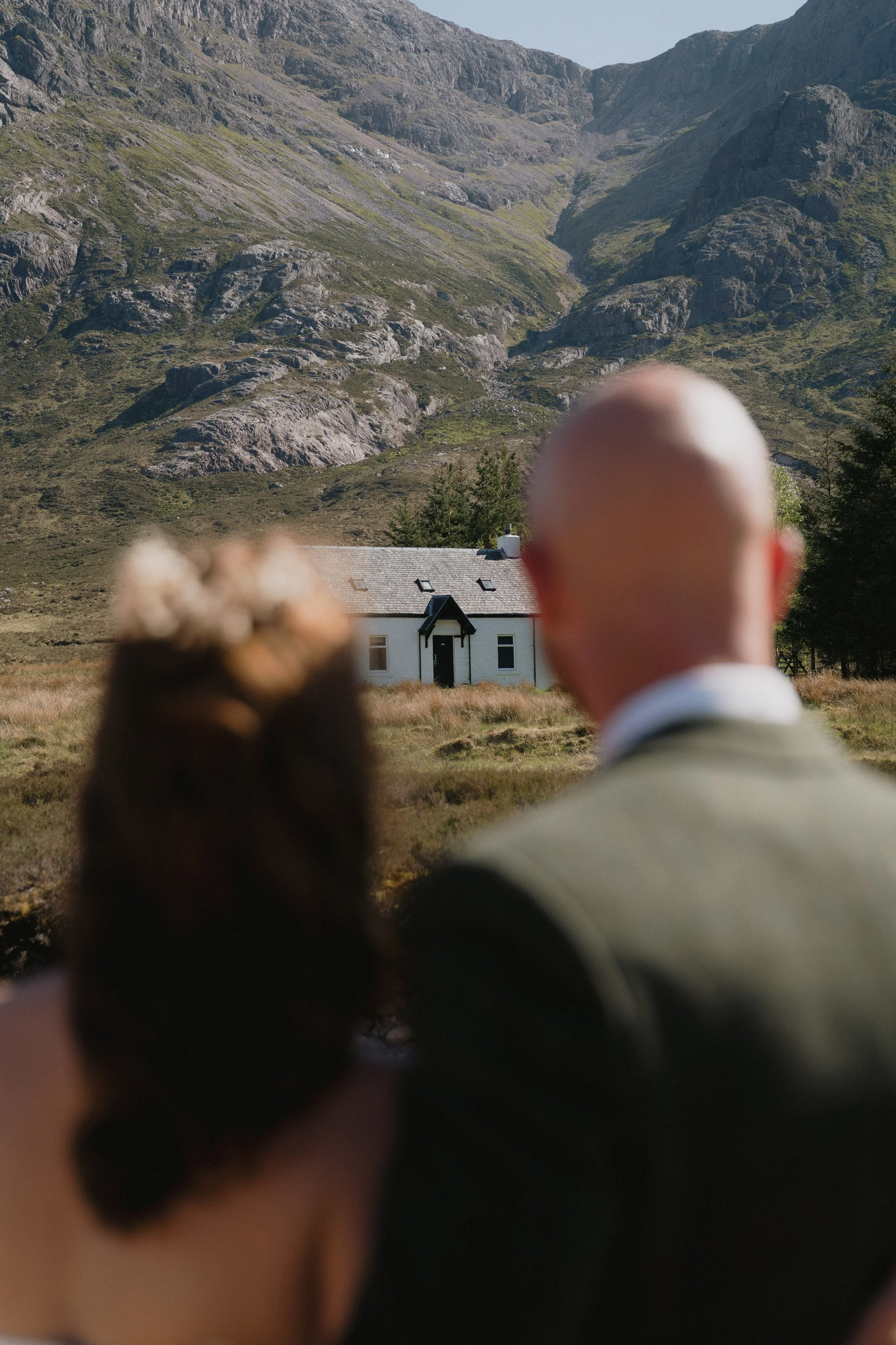 A couple, a woman with flowers in her hair and a bald man, are standing outdoors on a grassy area, looking at a white house with a black roof, set against a backdrop of green mountains. - captured by an Edinburgh wedding photographer