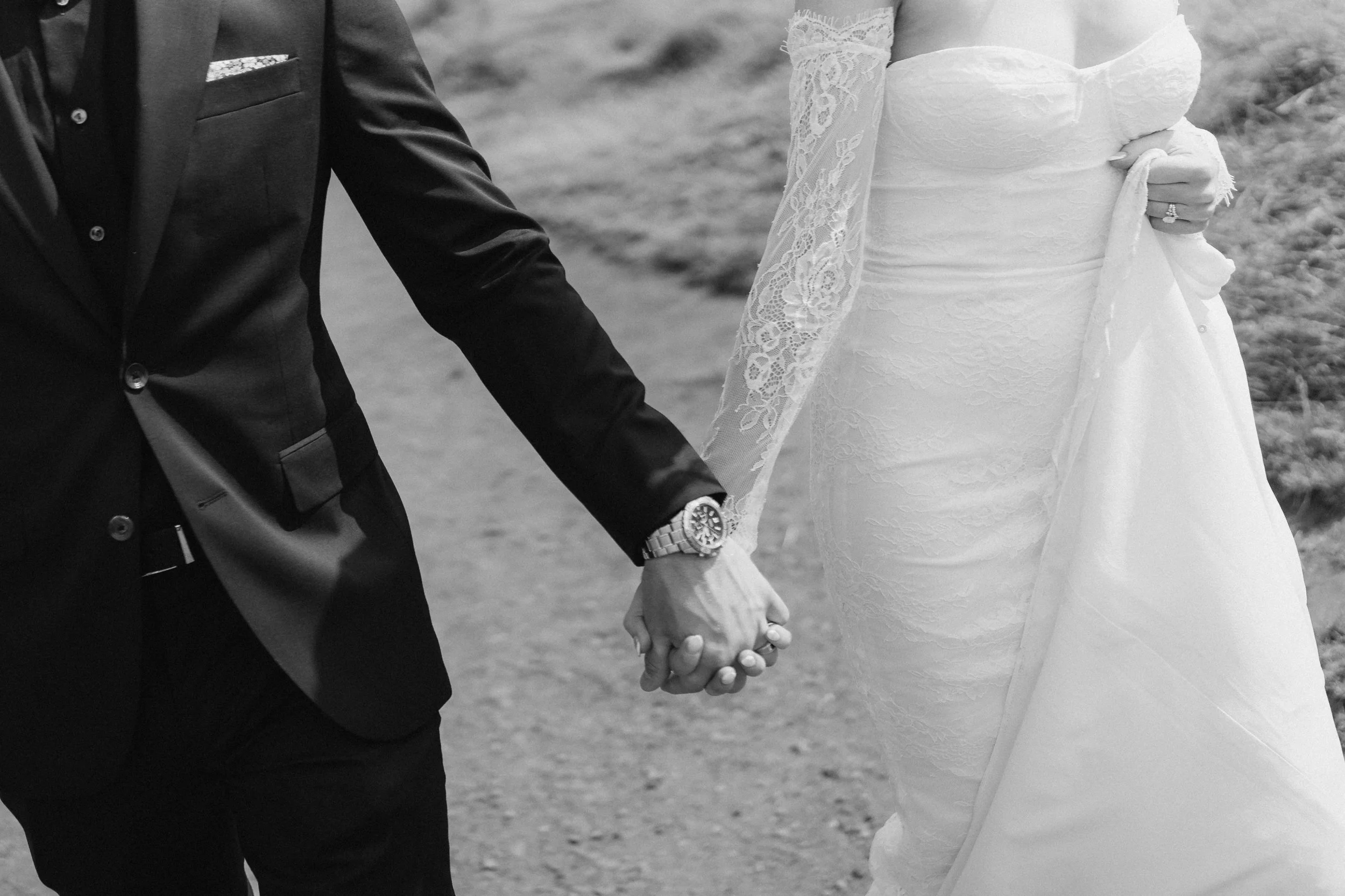 A black and white photo of a bride and groom holding hands during their wedding, with part of the bride's wedding dress and the groom's suit visible. - captured by an Edinburgh wedding photographer