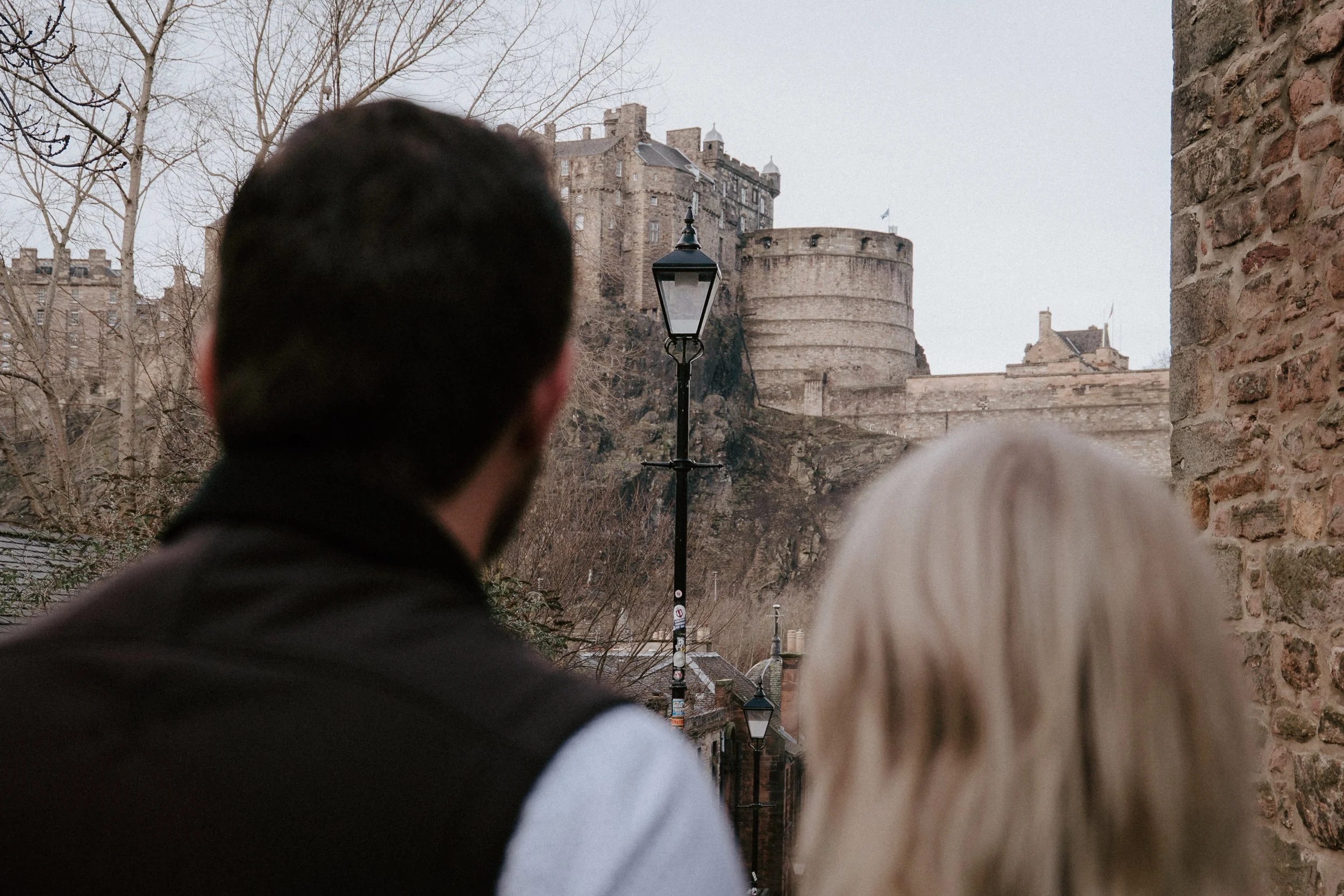Two people, a man with dark hair and a woman with blonde hair, are viewed from behind as they look at a historic castle on a hill, with leafless trees and brick walls in the foreground and a street lamp between them.