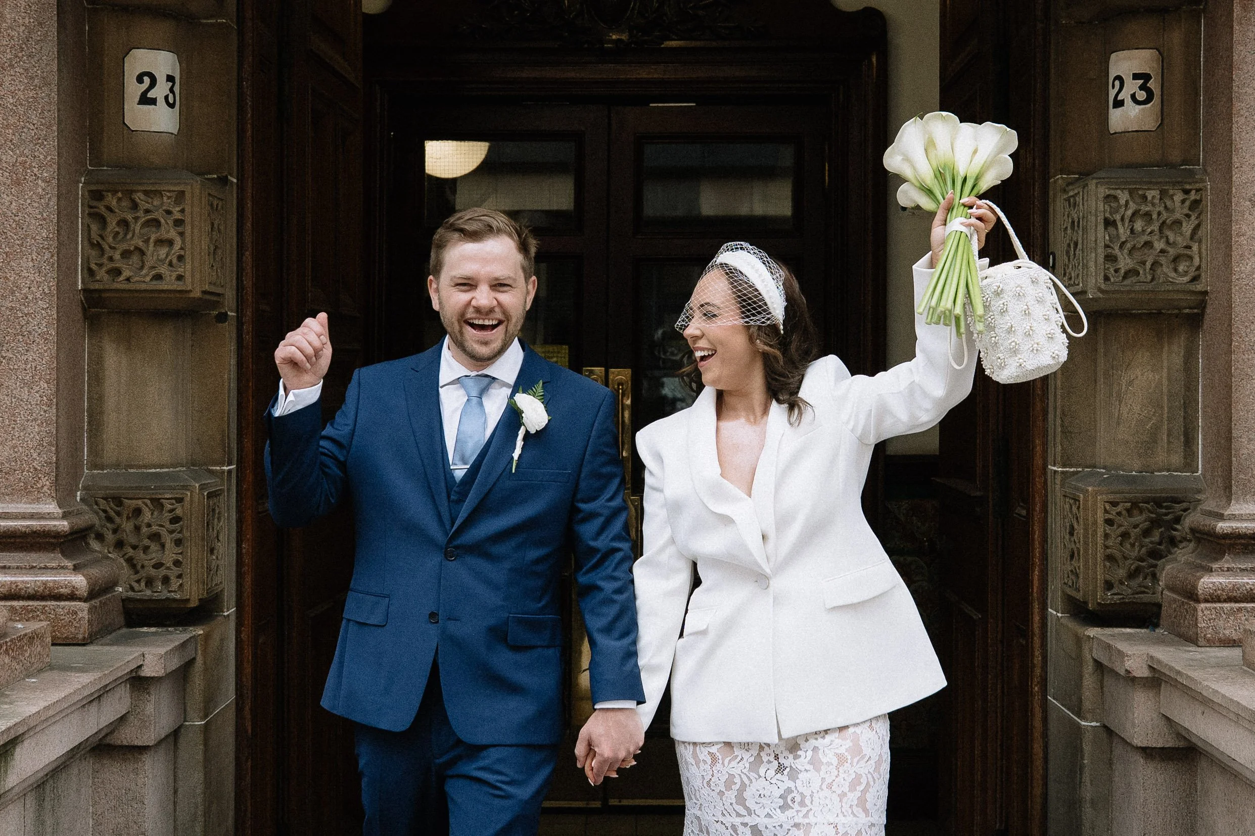 A newly married couple walking hand in hand out of a building, smiling and celebrating. The groom is wearing a blue suit with a white shirt and blue tie. The bride is dressed in a white lace dress with a white blazer and netted veil, holding a bouque