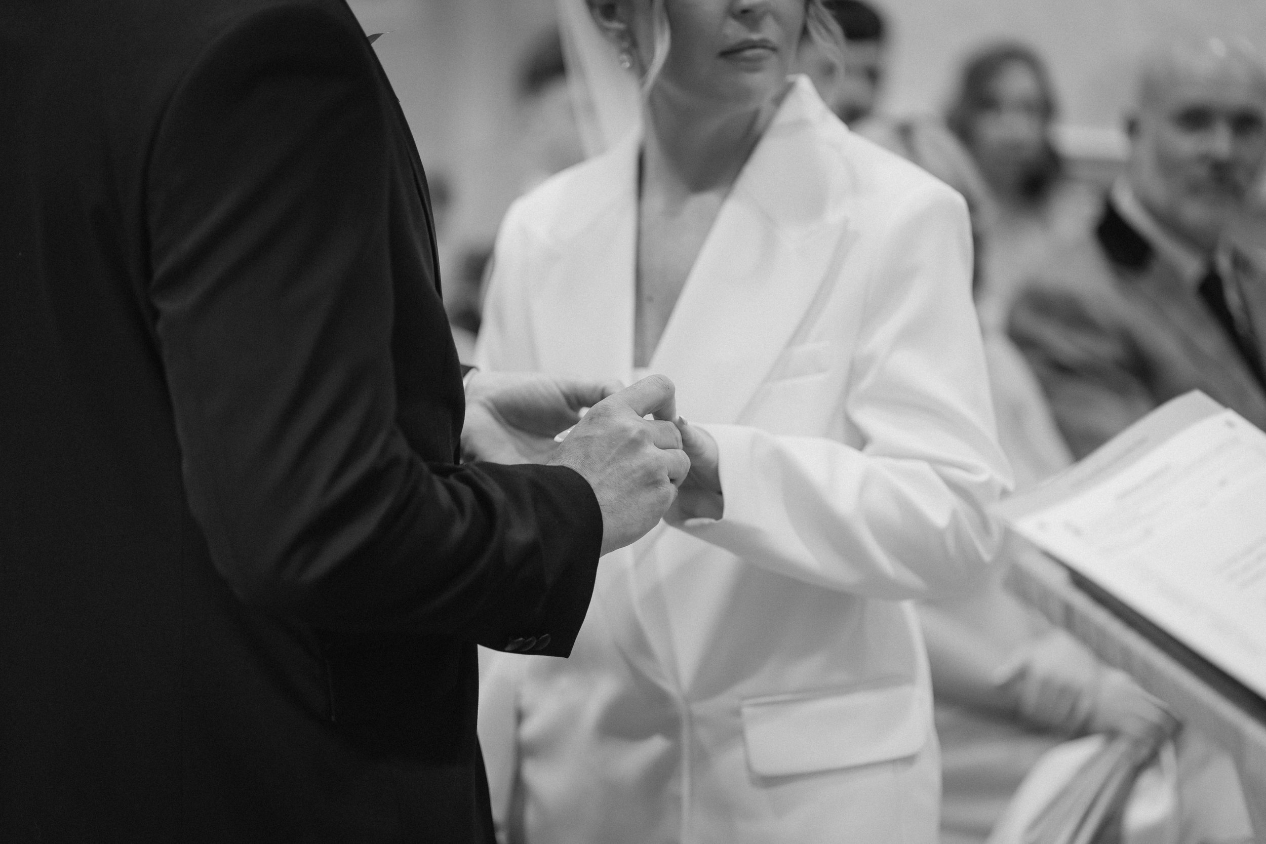 A person in a dark suit is holding hands with a woman in a white blazer during a formal event, with people seated and some wearing masks in the background. - captured by an Edinburgh wedding photographer