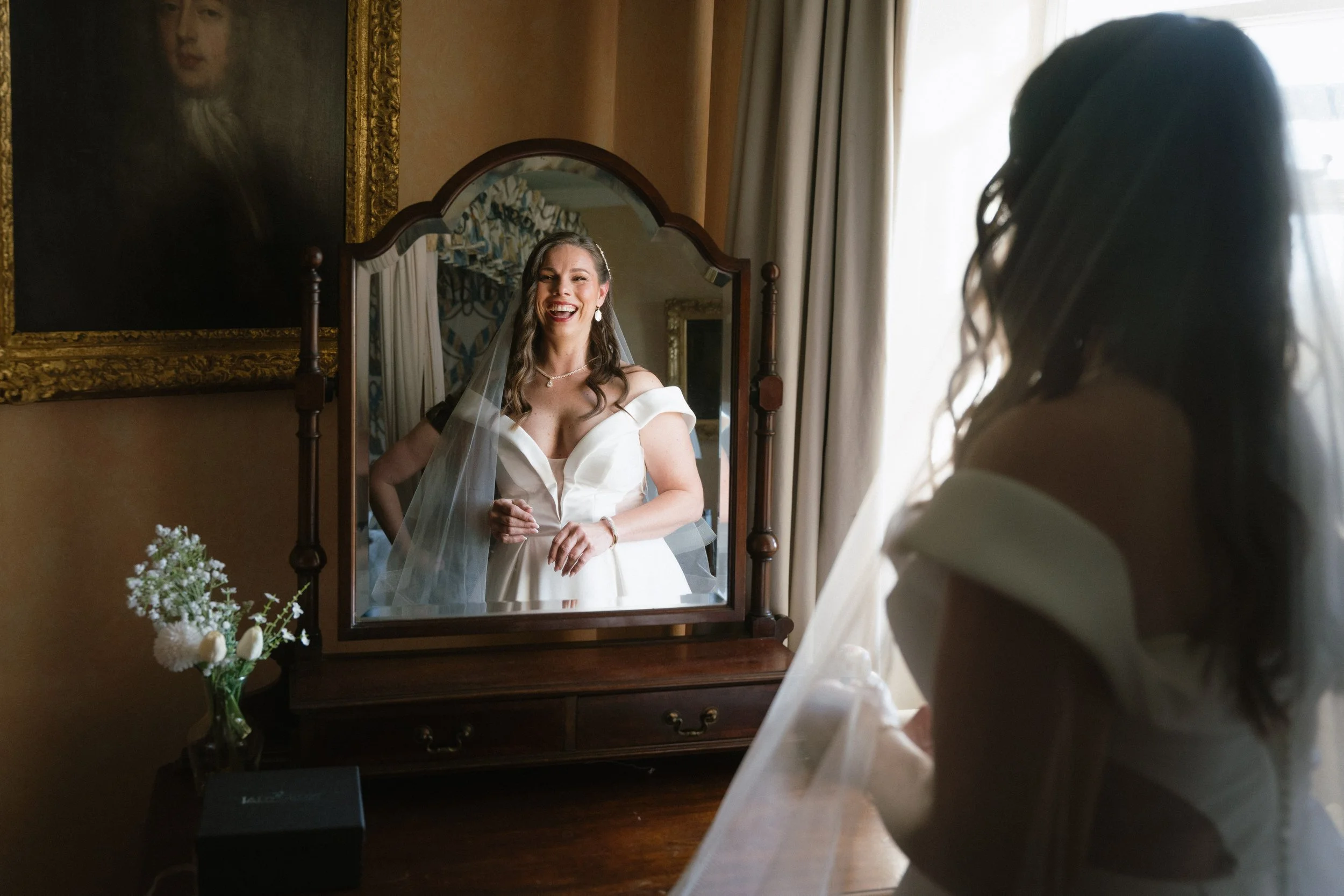 A bride in a white dress and veil smiling and looking at herself in a mirror. - captured by an Edinburgh wedding photographer