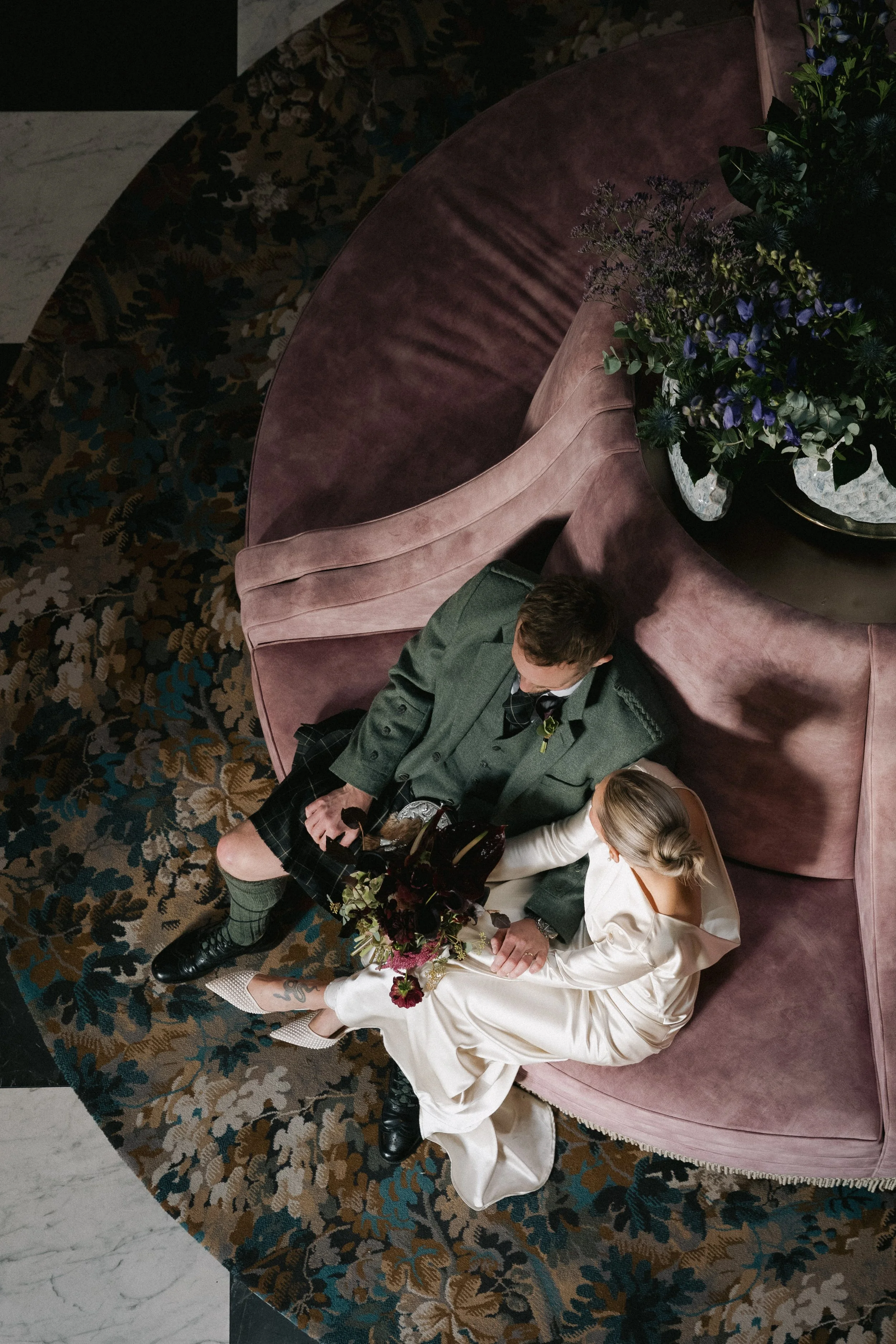 A man and woman sitting on a curved mauve velvet sofa, viewed from above. The woman is dressed in a cream-colored satin dress with long sleeves, holding a bouquet of flowers. The man is wearing a traditional Scottish kilt and jacket. They are surroun