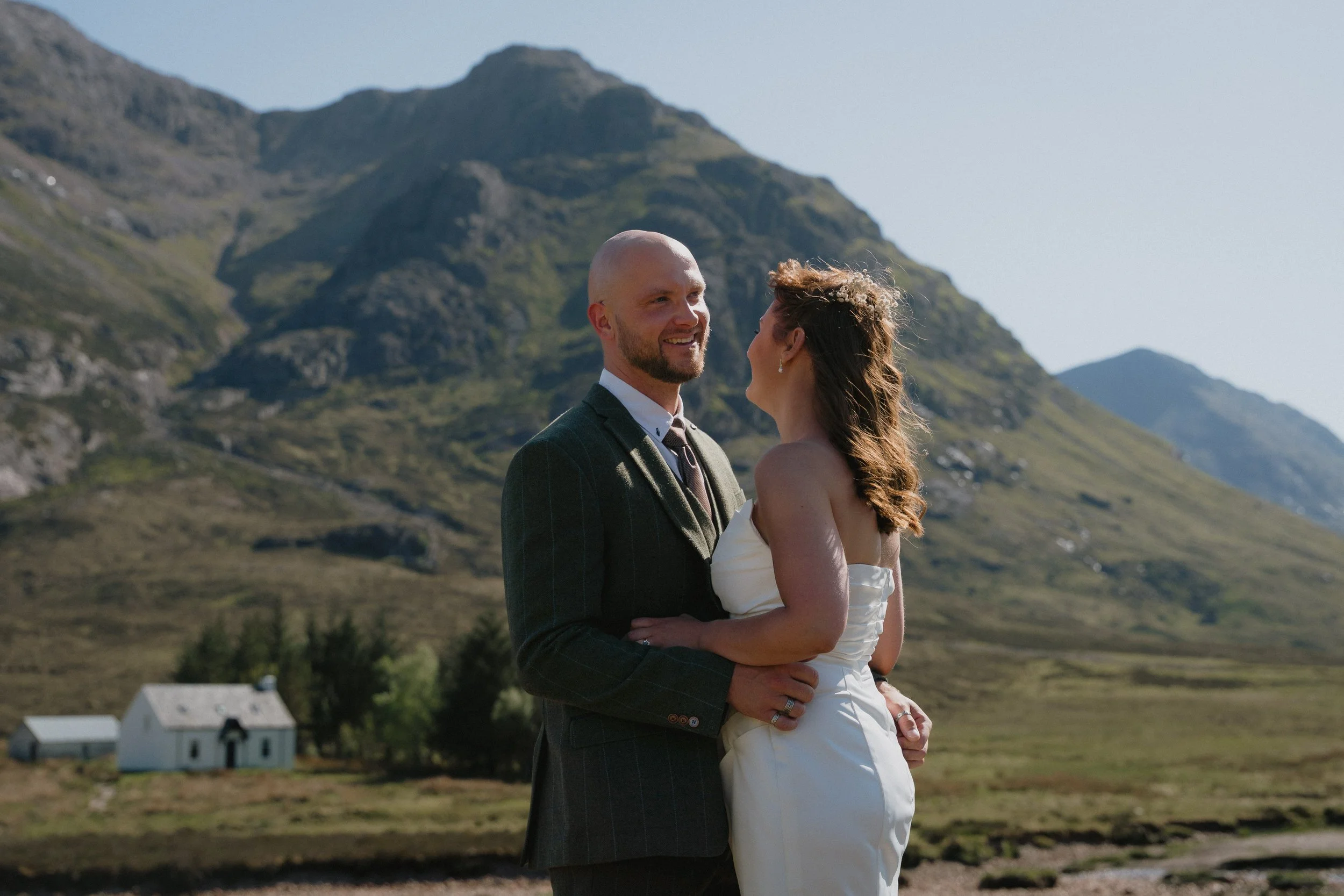 A newlywed couple embracing outdoors with mountains in the background, a small white church and greenery around. - captured by an Edinburgh wedding photographer