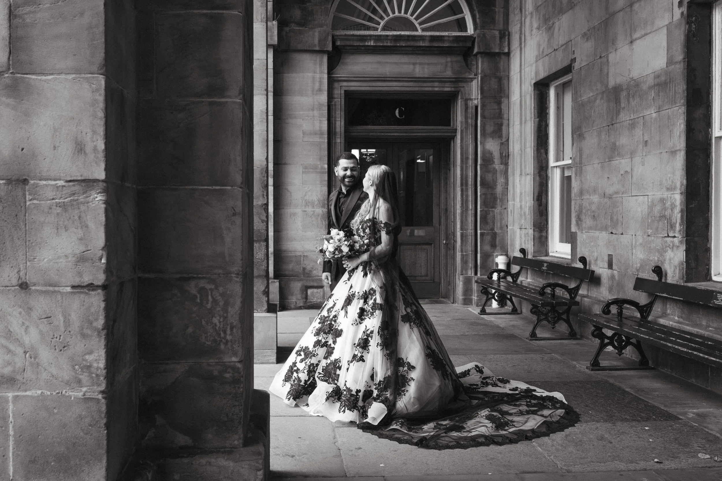 A bride and groom standing close together, smiling, in front of a historic stone building. The bride is wearing a floral wedding gown and holding a bouquet, while the groom is in a dark suit. They are under a stone archway with benches and windows ne